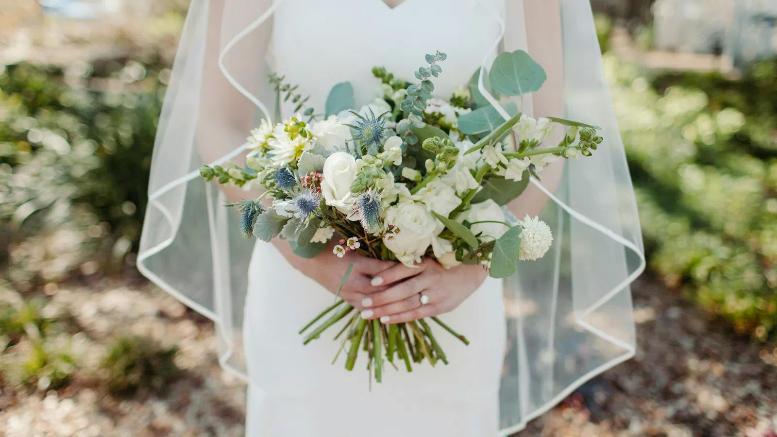 Bride in white dress holding bouquet of white and green flowers with veil outdoors on sunny day