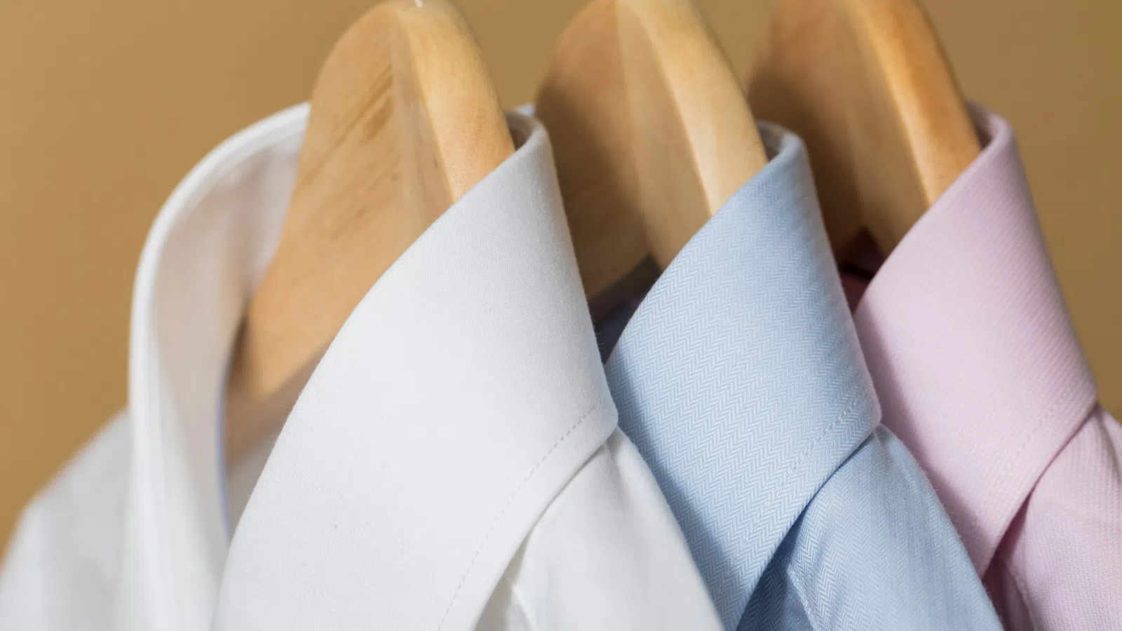 White, blue, and pink dress shirts neatly hanging on wooden hangers against a beige background