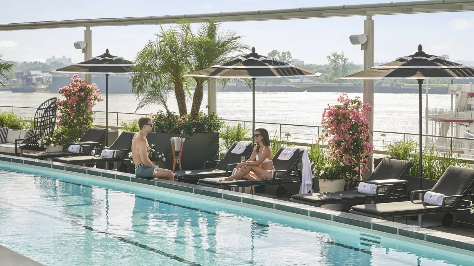 Couple relaxing and talking on lounge chairs by a pool with umbrellas and river view in the background
