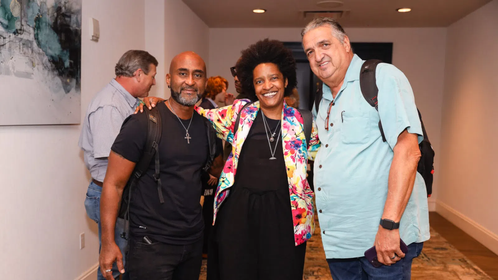Three diverse adults smiling and posing together indoors at a social event in a hallway.