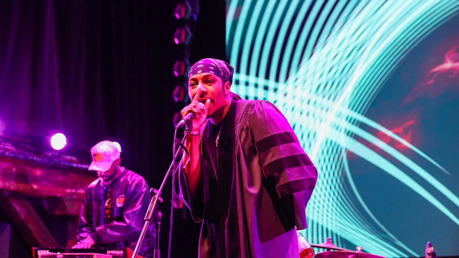 Hip-hop artist performing on stage with microphone and DJ in the background under vibrant purple and blue lights.