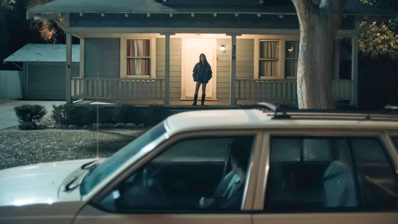 Person stands on lit porch of house at night with car parked in driveway in the foreground.
