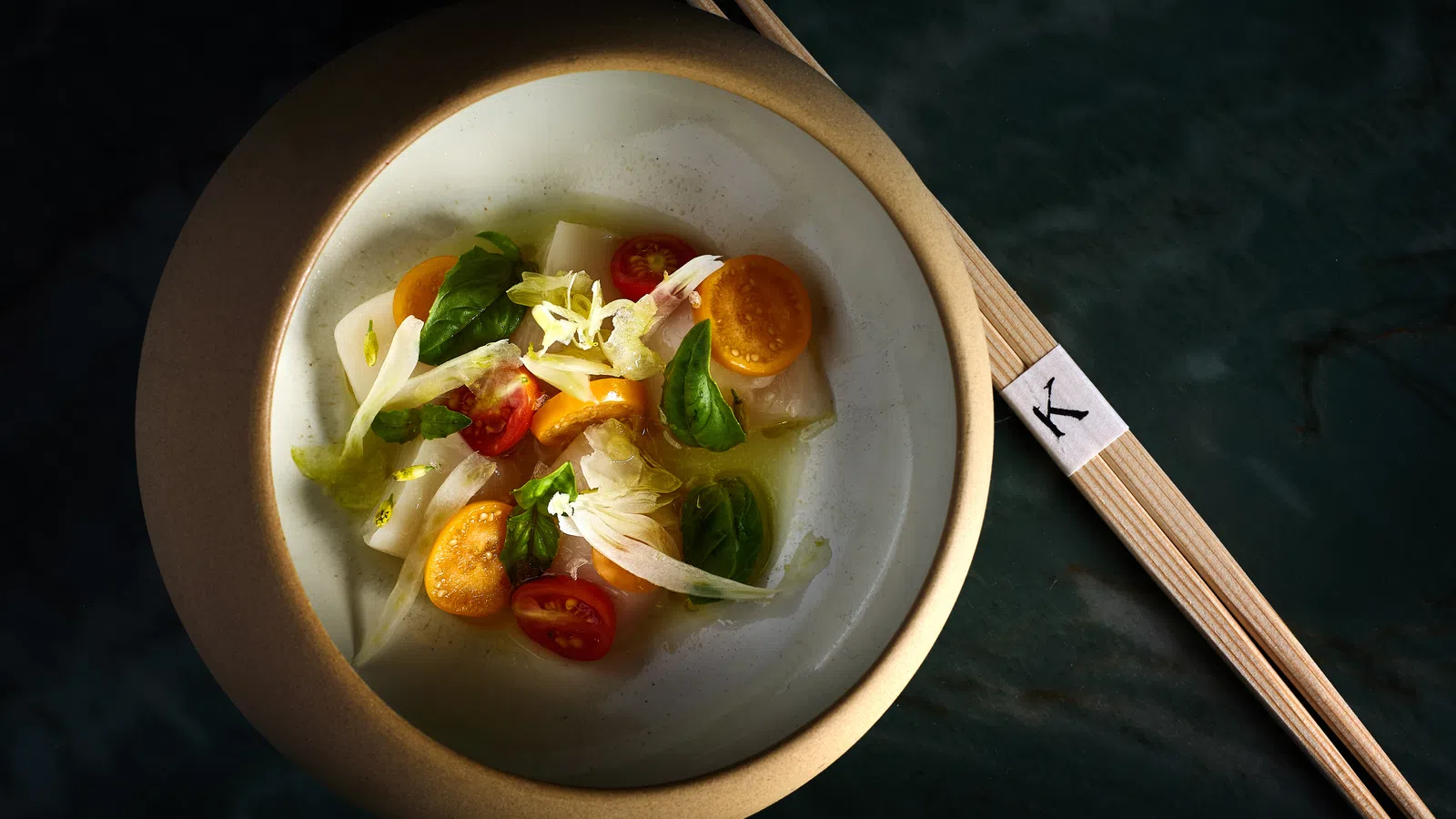 Elegant bowl of fresh vegetable salad with cherry tomatoes, greens, and chopsticks on dark surface.