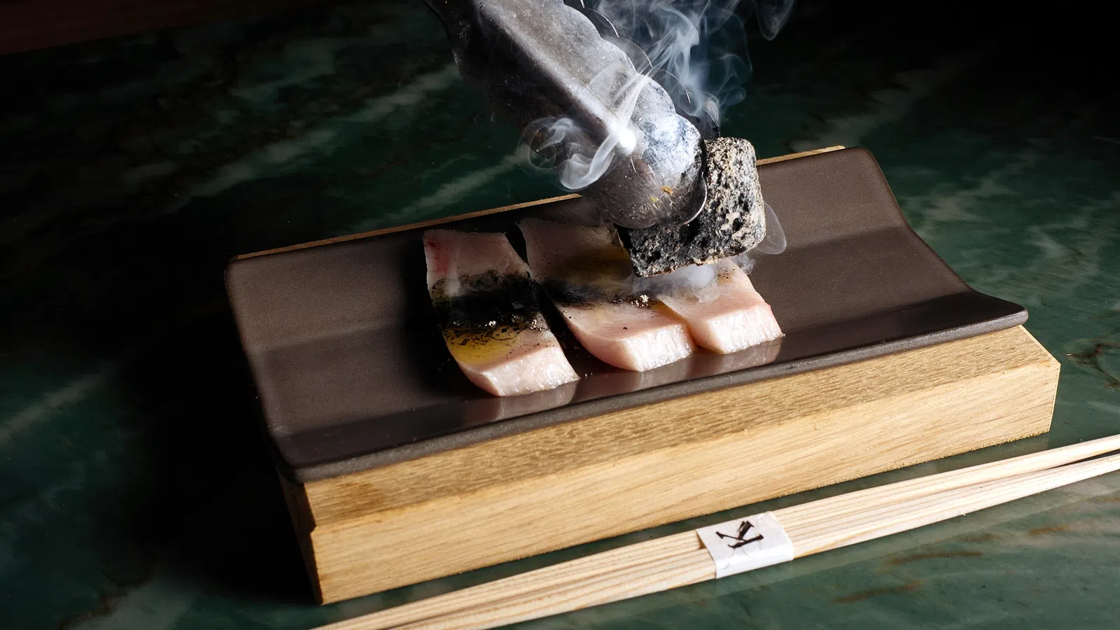 Close-up of sizzling fish slices being grilled with smoking charcoal on a rectangular plate with chopsticks.