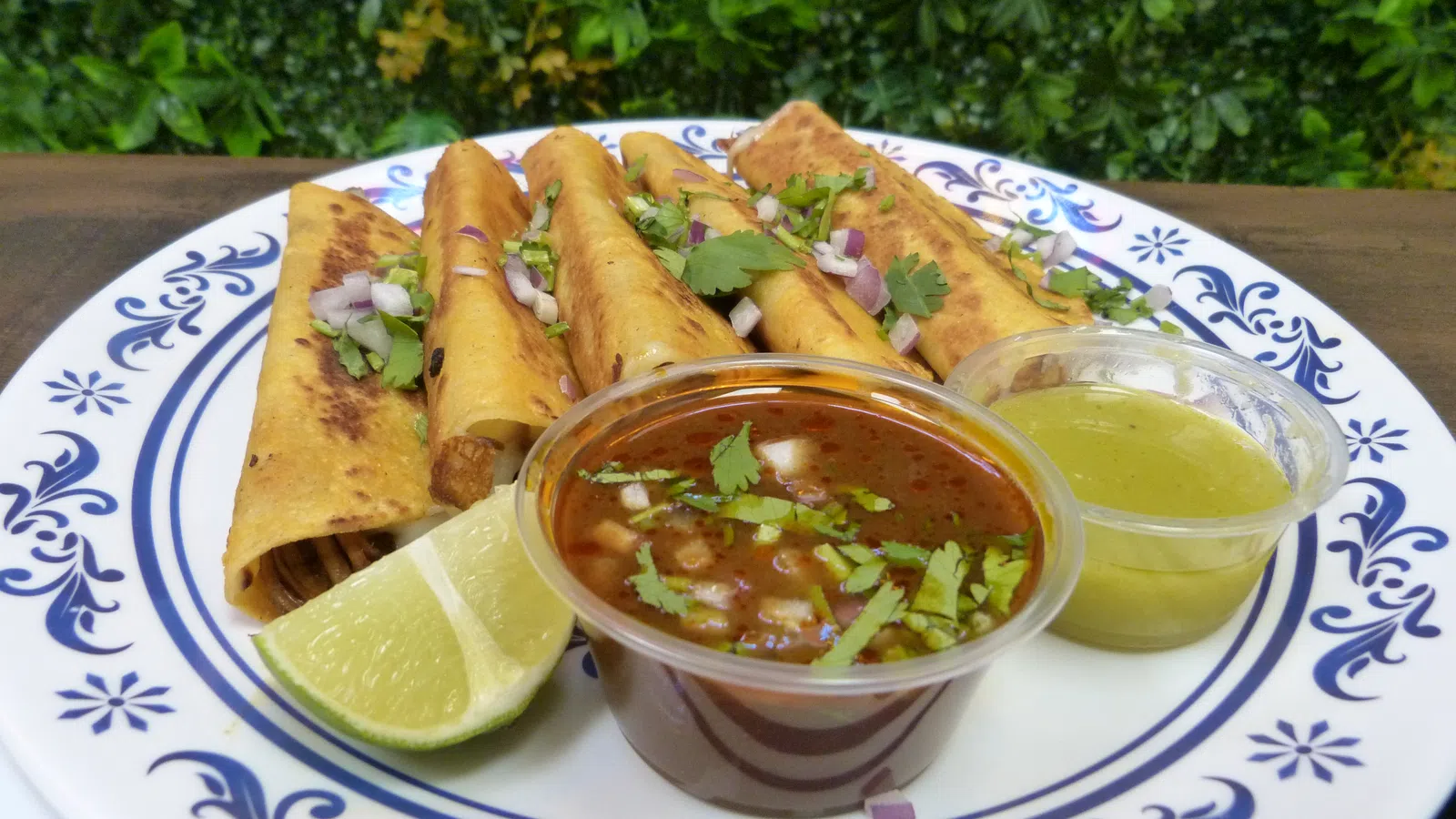 Plate of golden taquitos garnished with cilantro and onions, served with lime wedges and two dipping sauces.