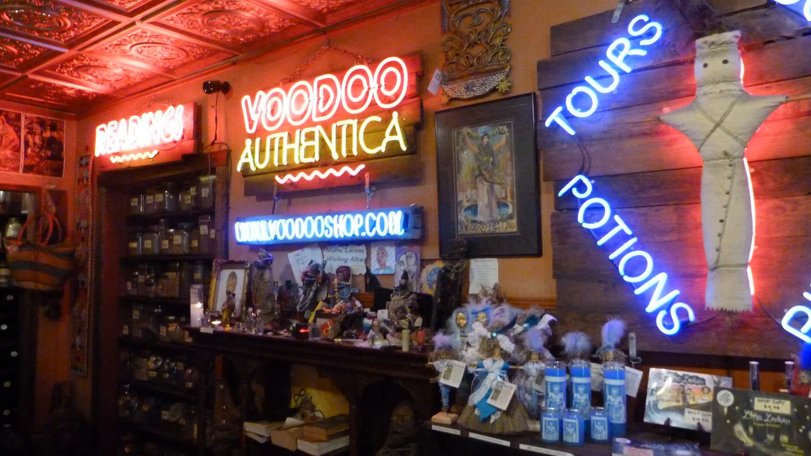 Interior of a voodoo shop with neon signs, shelves of jars, potions, dolls, and mystical items under ornate ceiling tiles.
