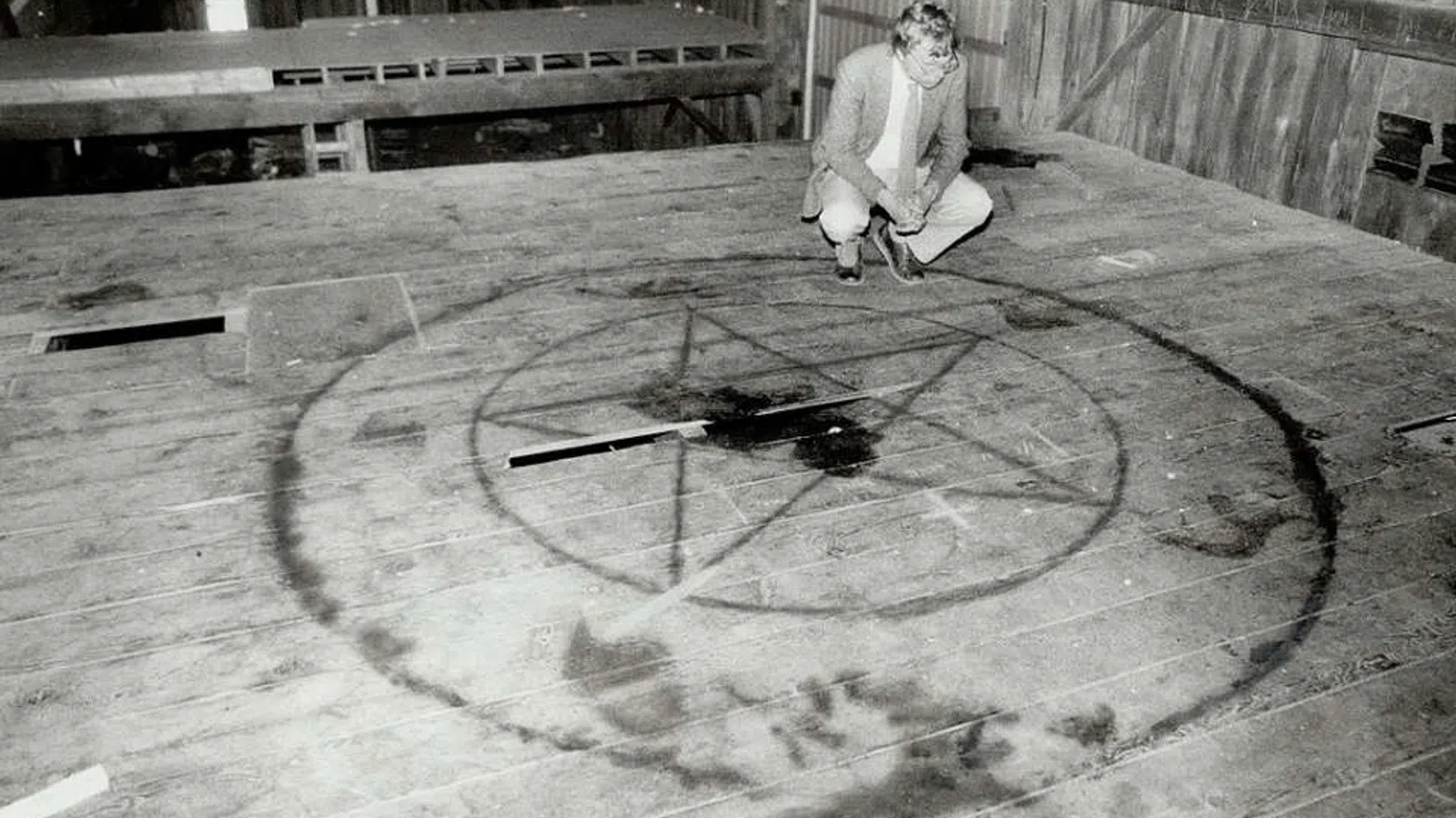 Black and white image of a man inspecting a large pentagram symbol drawn on a wooden floor inside a barn.