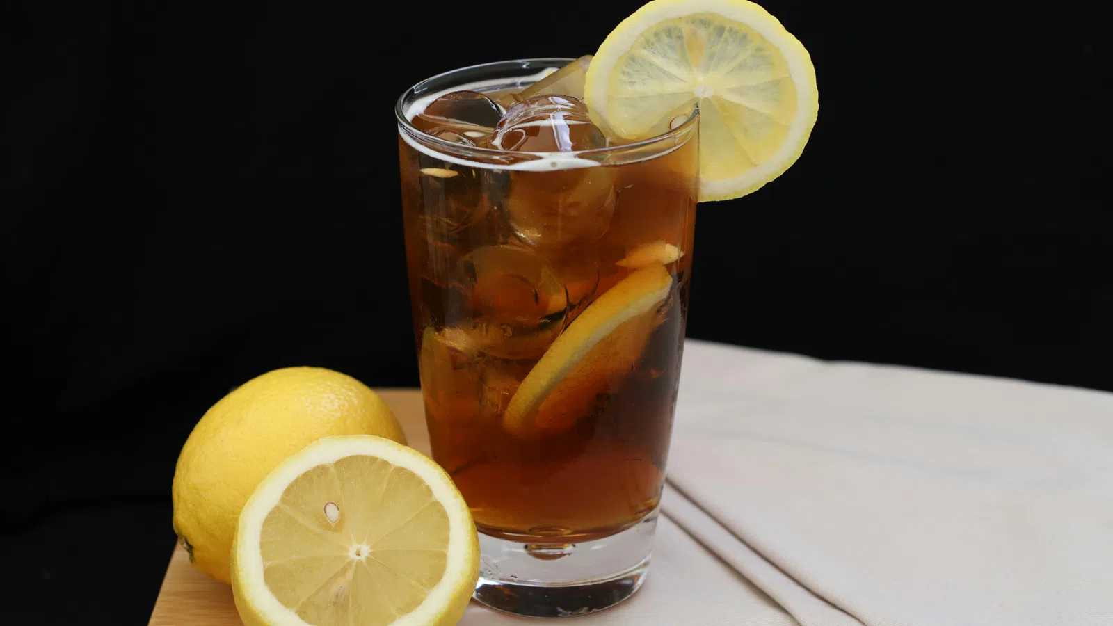 Glass of iced tea with lemon slices and whole lemons on wooden surface against black background.