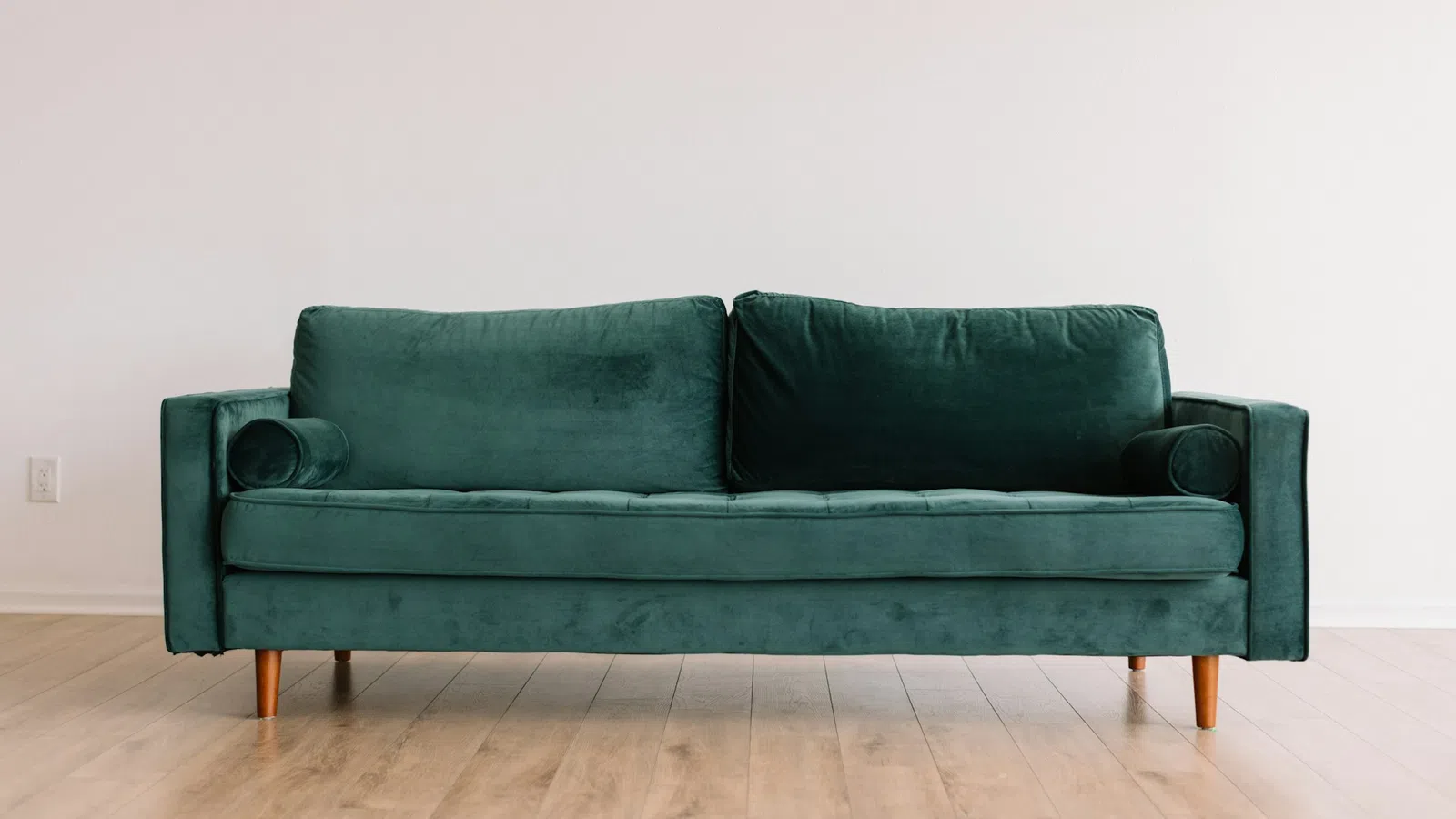 Modern emerald green velvet sofa with wooden legs on light wooden floor against plain white wall