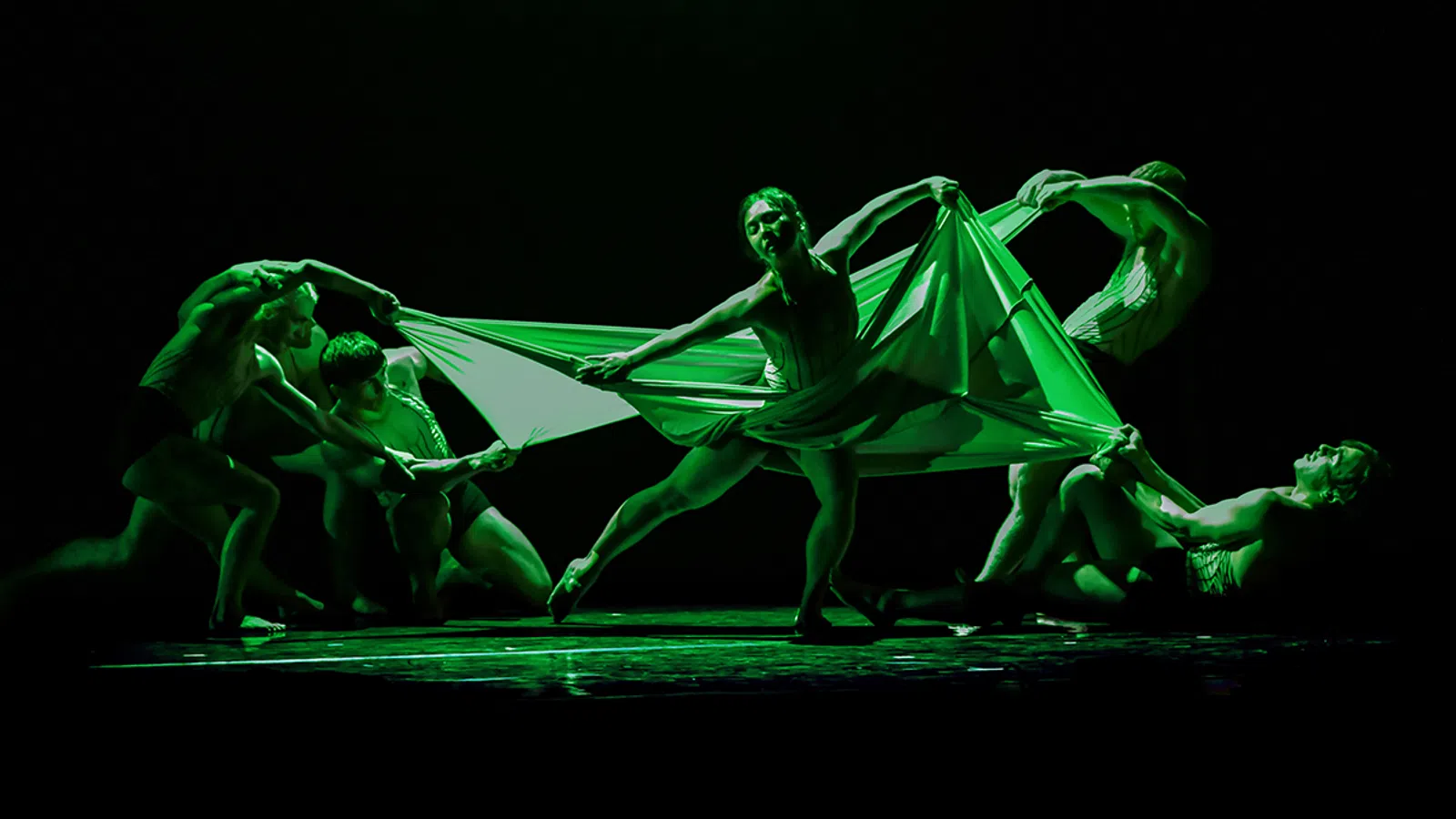 Performers in a dramatic green-lit dance holding and stretching a large fabric on stage in dynamic poses.