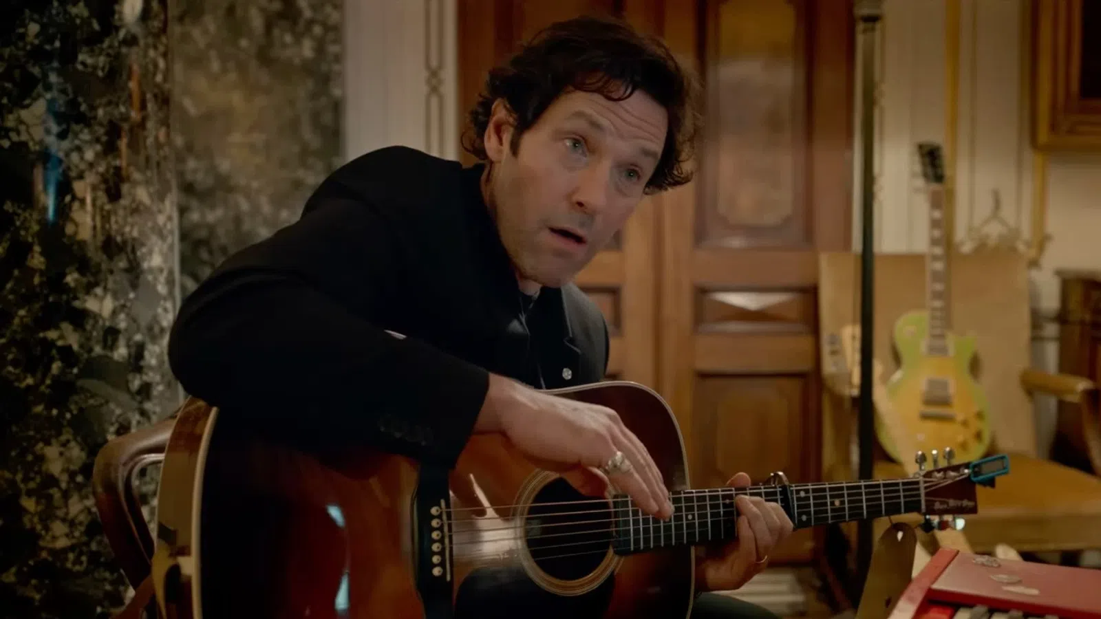 Man in black jacket playing acoustic guitar indoors with wooden doors and electric guitar in background