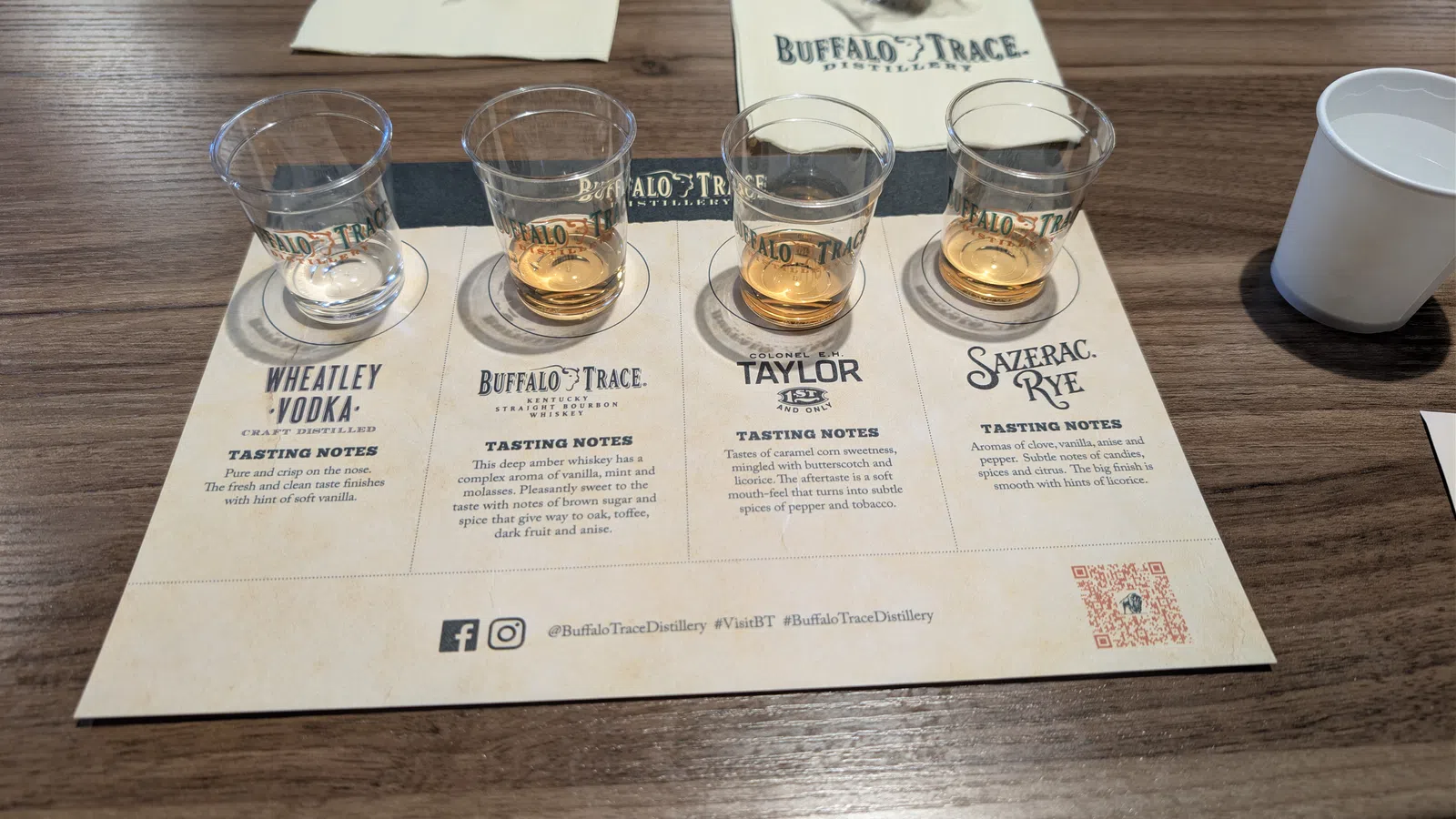 Four whisky tasting samples on a Buffalo Trace Distillery flight tasting mat with tasting notes and a cup on a wooden table.