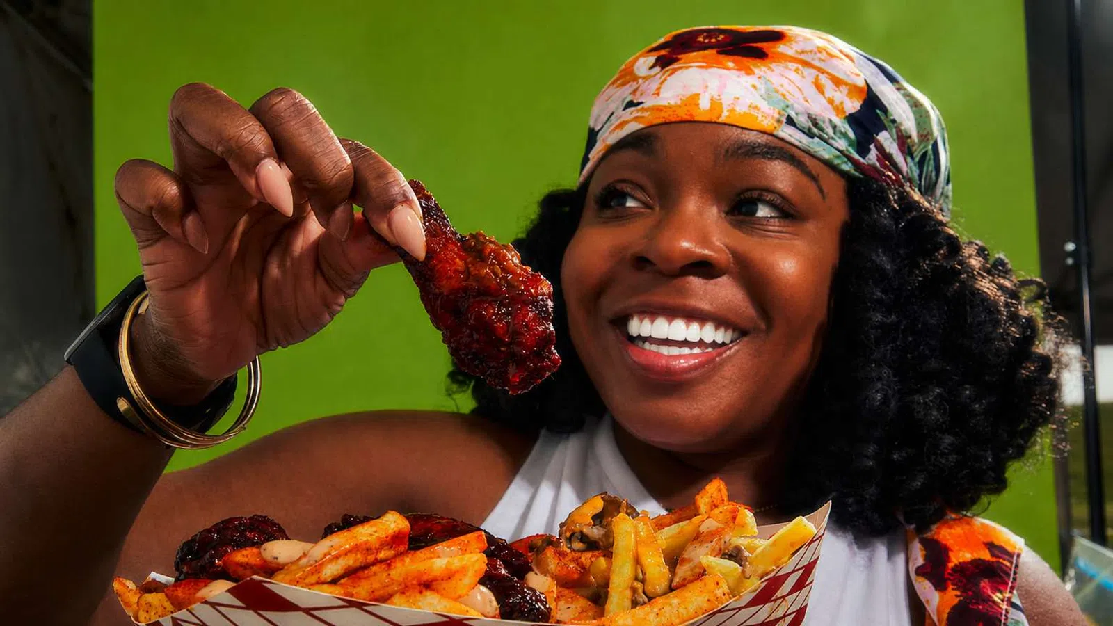 Smiling woman holding a saucy chicken wing and a tray of seasoned fries against a green background.