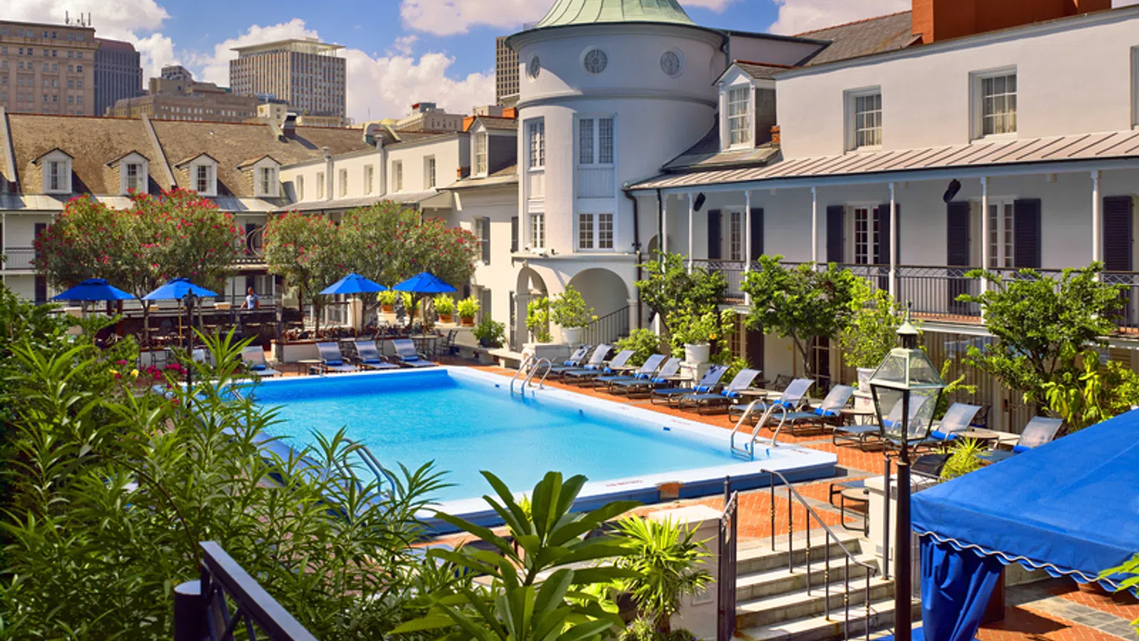 Outdoor pool area surrounded by lounge chairs, greenery, and historic white buildings under a blue sky.