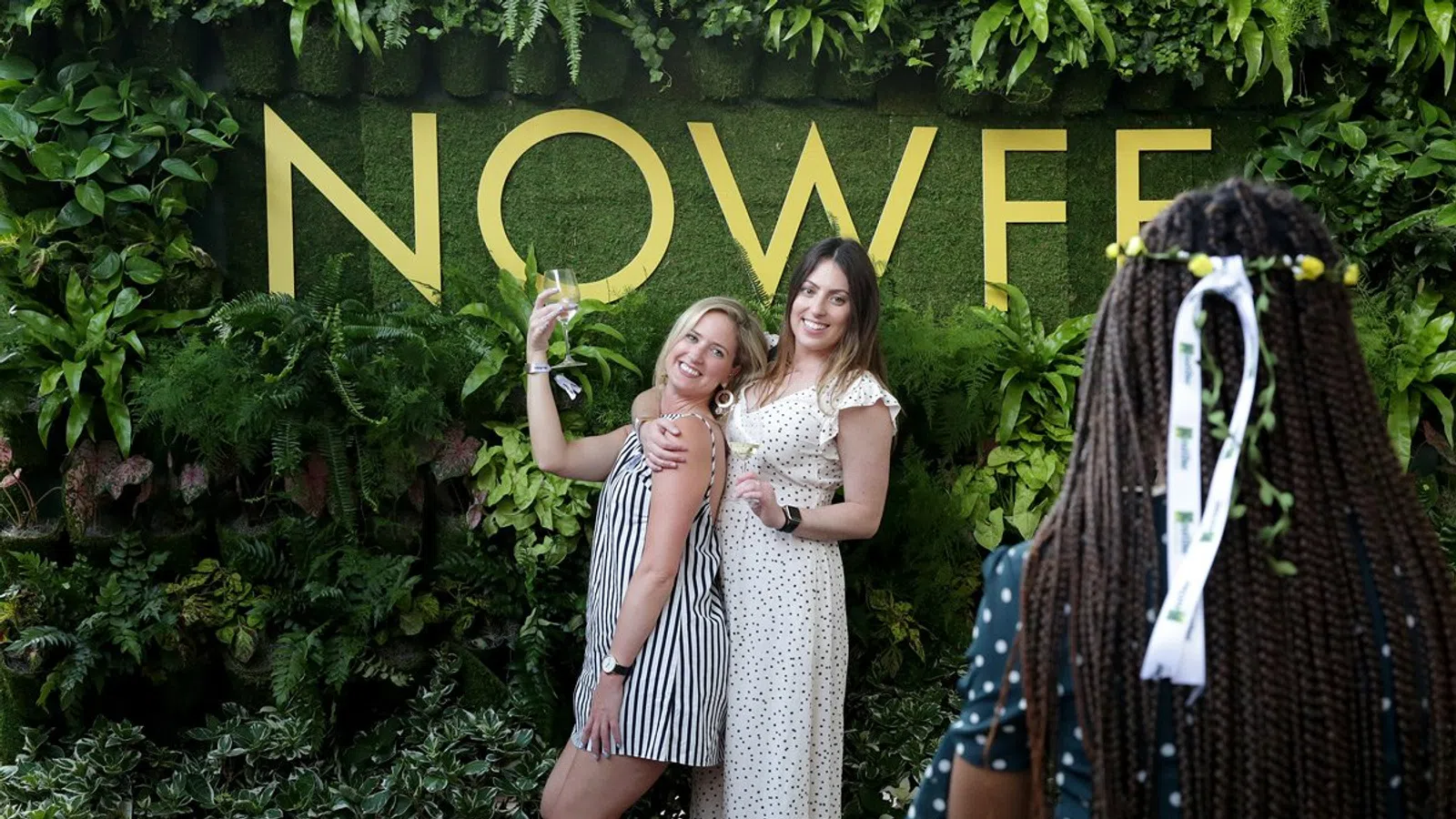 Two women pose and smile in front of a green plant wall with large yellow NOWEF letters while another person photographs them.