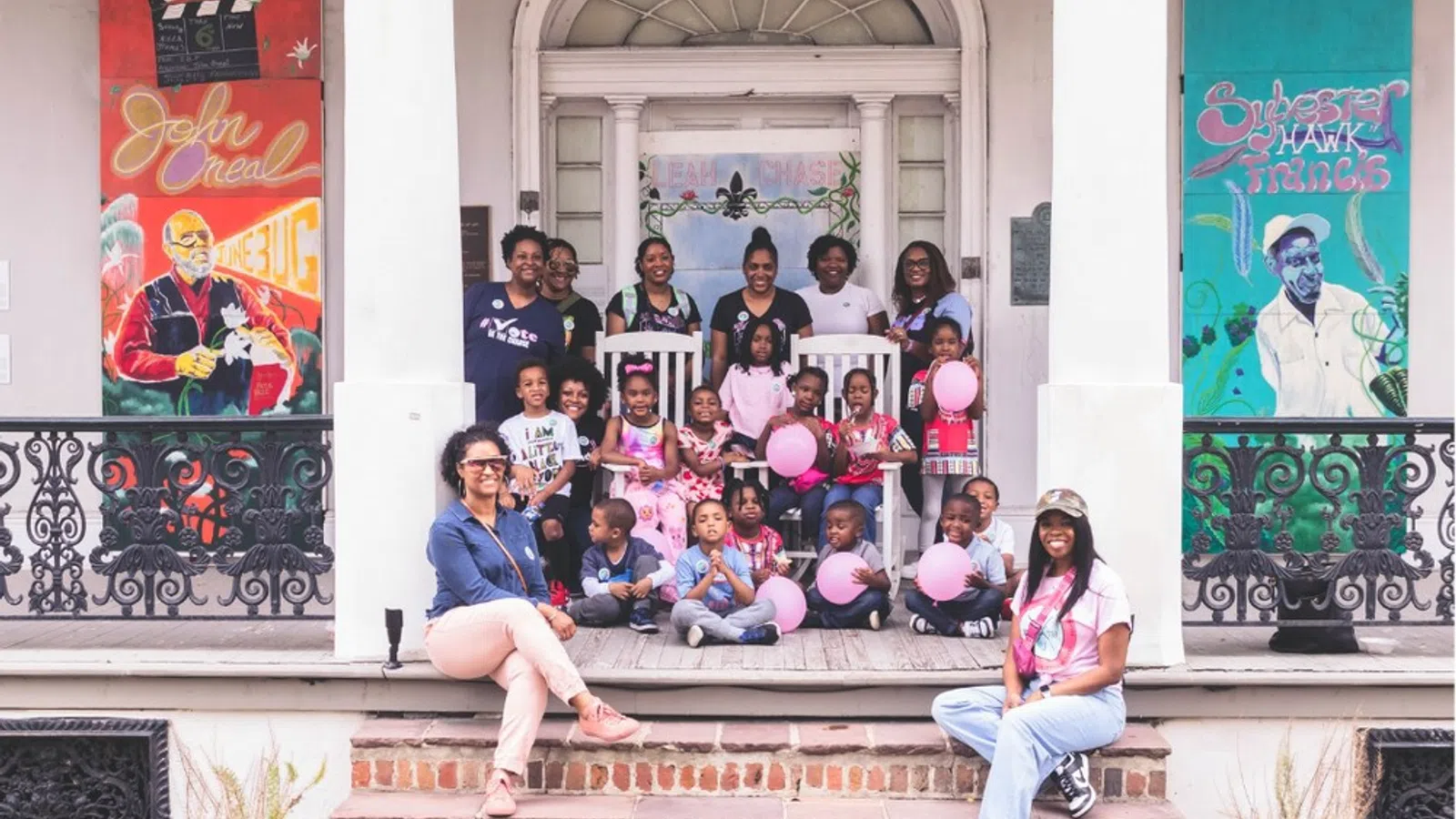 Group of adults and children posing with pink balloons on porch with colorful artwork on walls