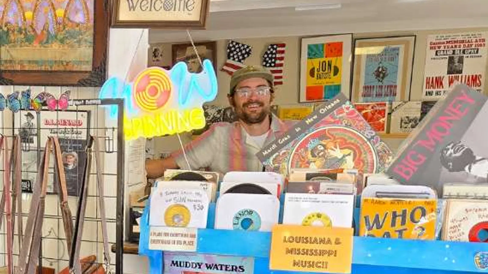 Smiling man behind record store counter with vinyl records, vintage posters, and neon open sign
