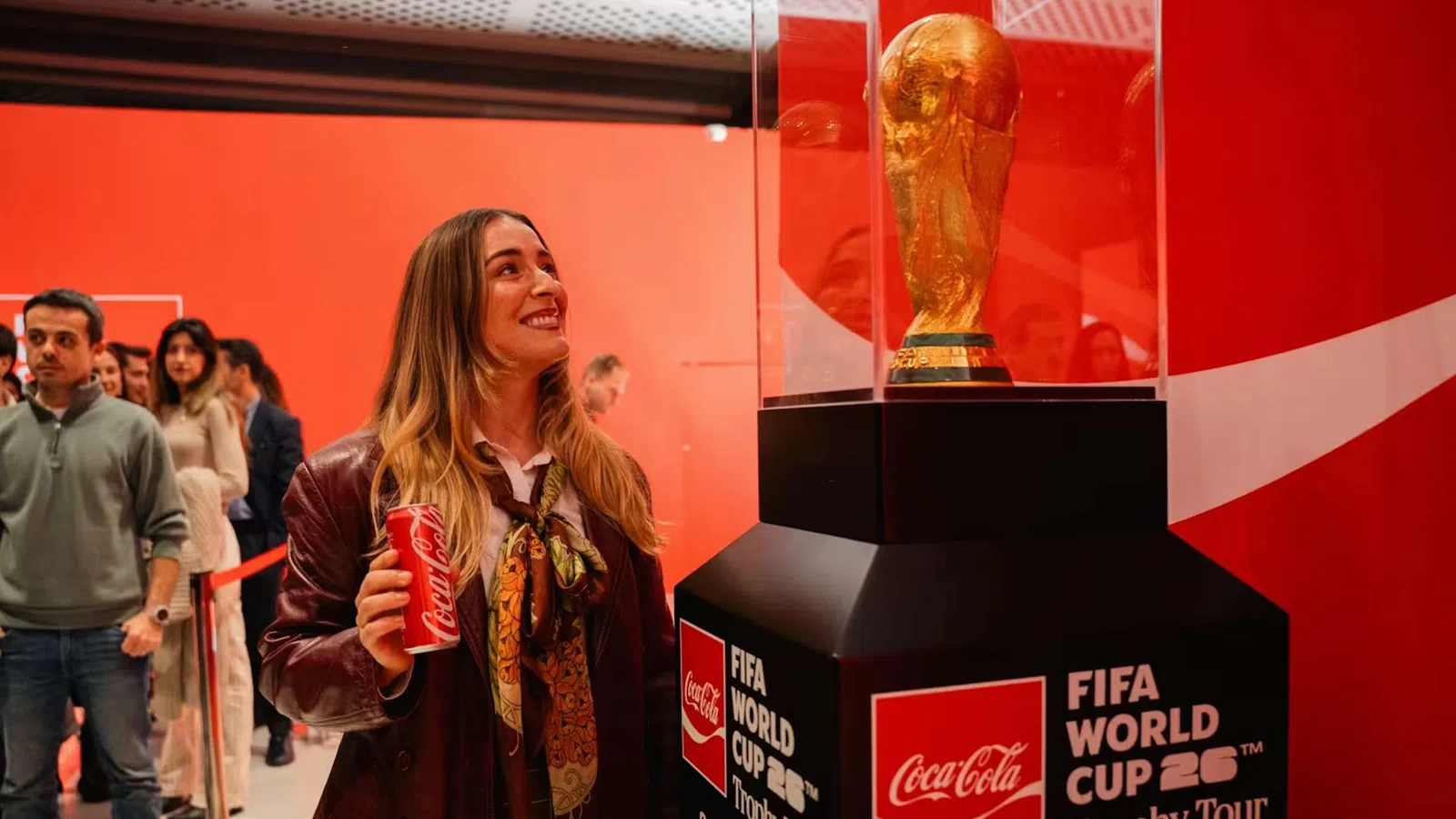 Woman holding Coca-Cola can looks at FIFA World Cup trophy in red-themed exhibit during trophy tour.