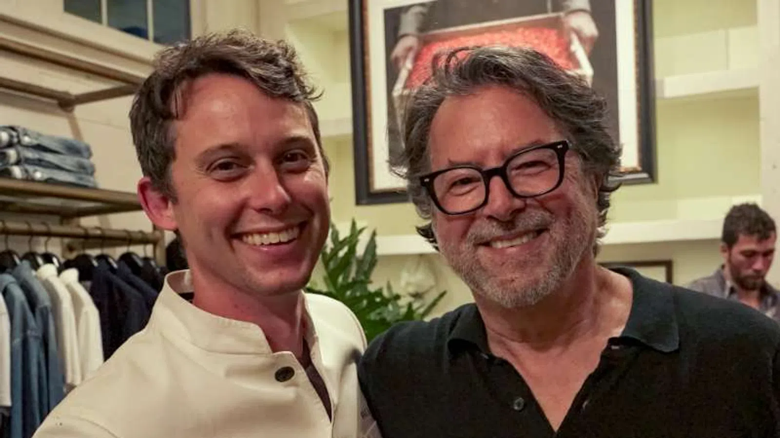 Two men smiling together indoors, one wearing a white chef jacket and the other in a black shirt with glasses.