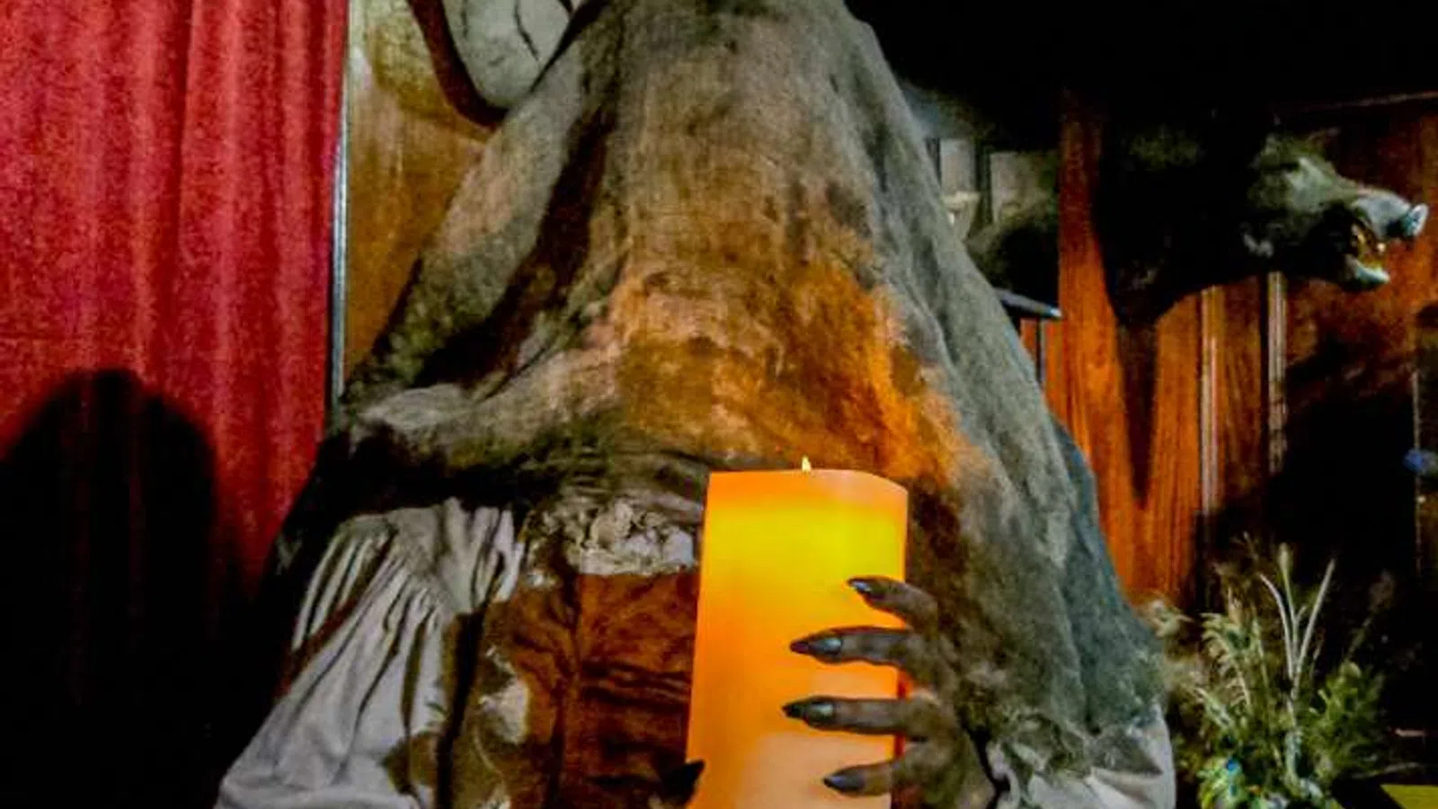 Ghostly figure with a cloth covering head holding a glowing candle in a dimly lit room with wooden walls.