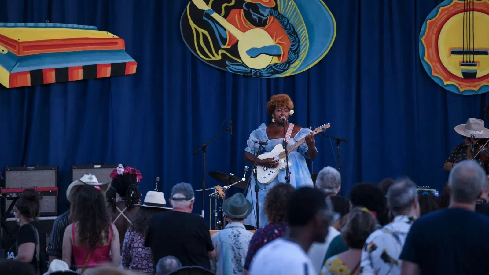 Singer with electric guitar performs on stage in front of crowd at outdoor music festival with blue backdrop.