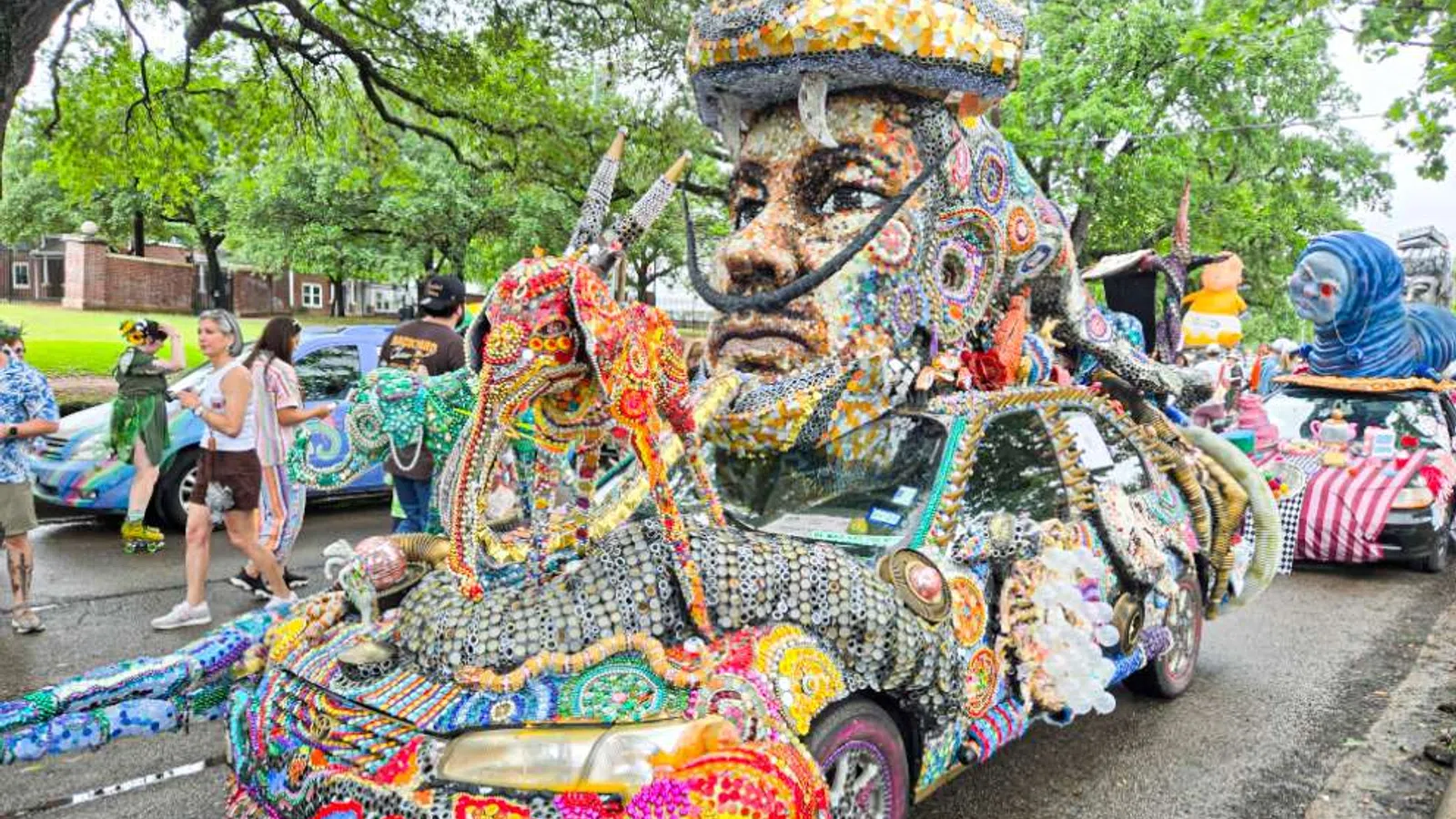 Colorful mosaic-covered car with a large head sculpture on top at an outdoor event with people and trees.