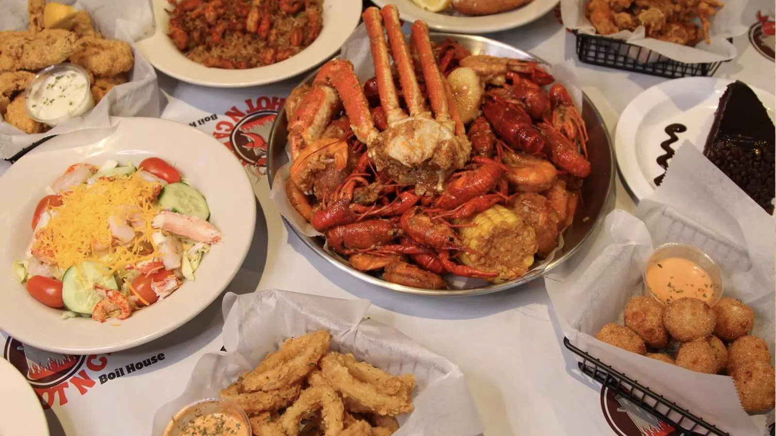 Seafood feast with crab, crawfish, fried shrimp, hush puppies, salad, and dessert on a white table.