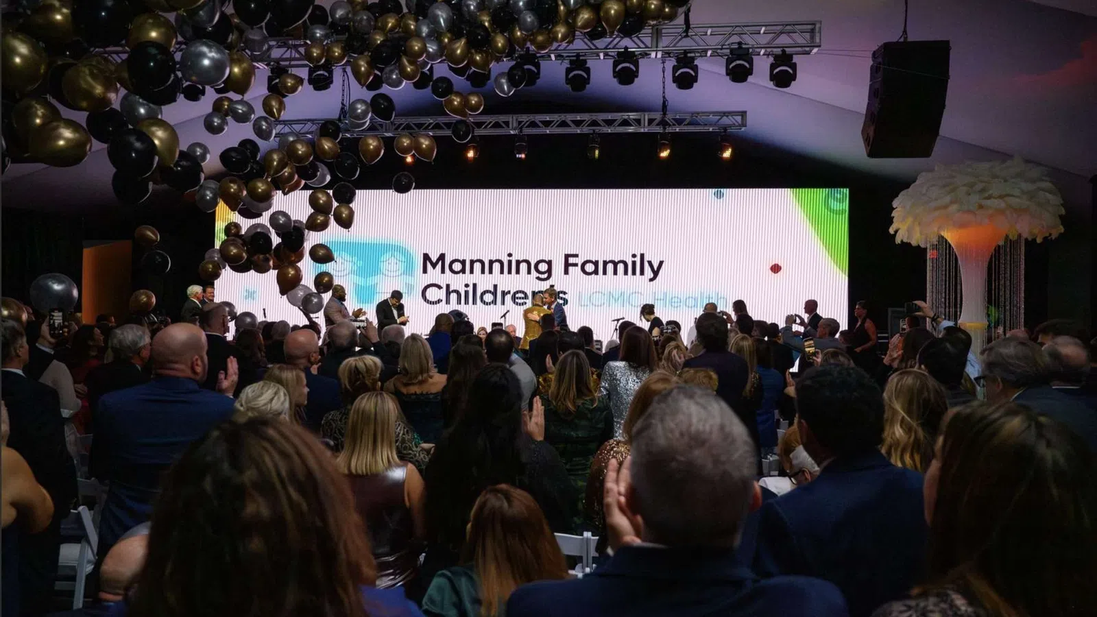 Audience applauding at a formal event with a large screen displaying Manning Family Children's LCMC Health