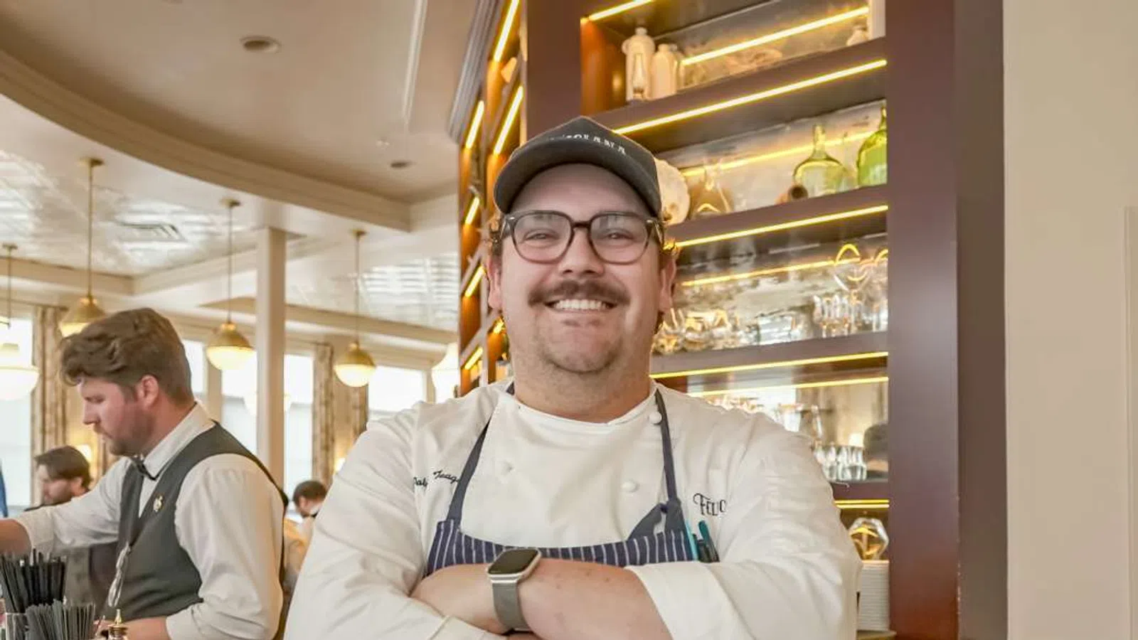 Smiling chef with glasses and cap stands with arms crossed in a restaurant kitchen with staff working in the background