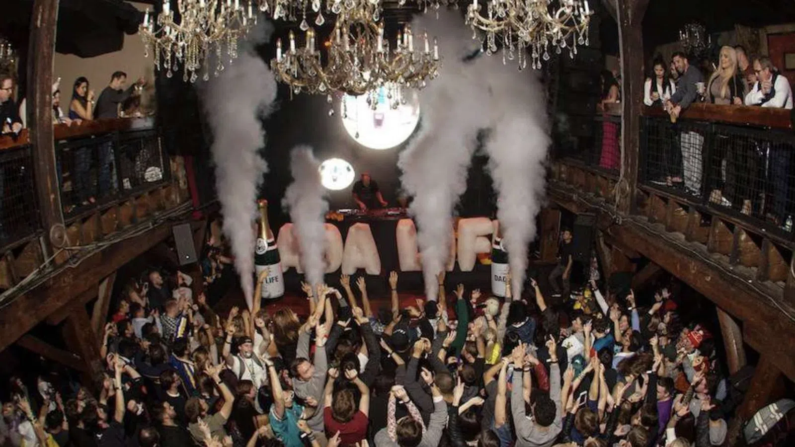 Crowded nightclub with people celebrating under chandeliers and large smoke effects from bottles on the dance floor.