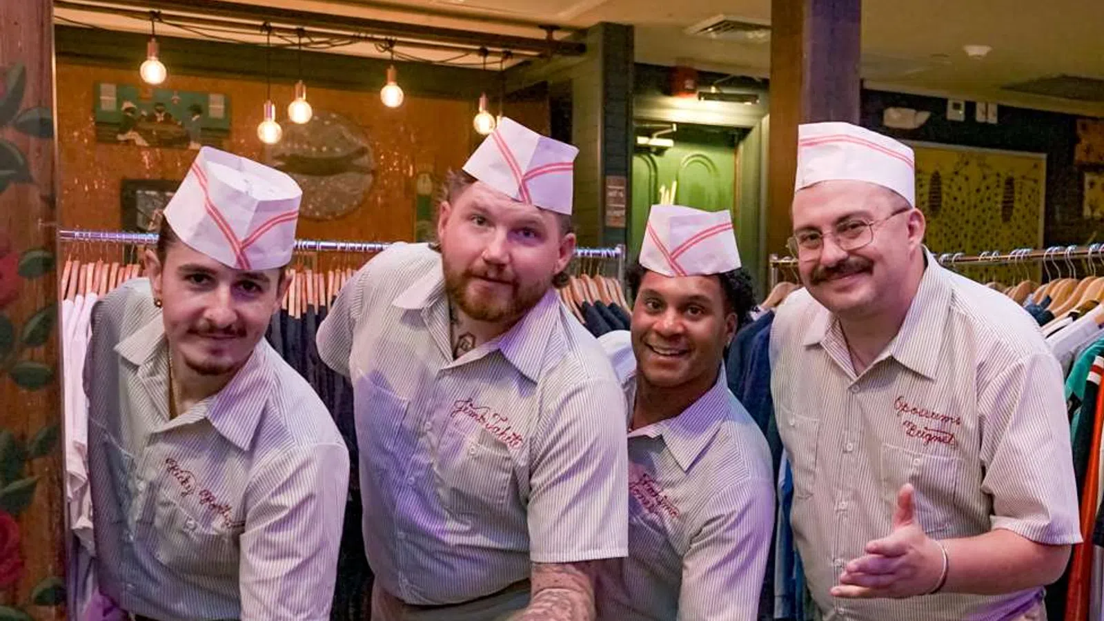 Four men wearing retro diner uniforms and hats posing with outstretched hands in a cozy restaurant setting.