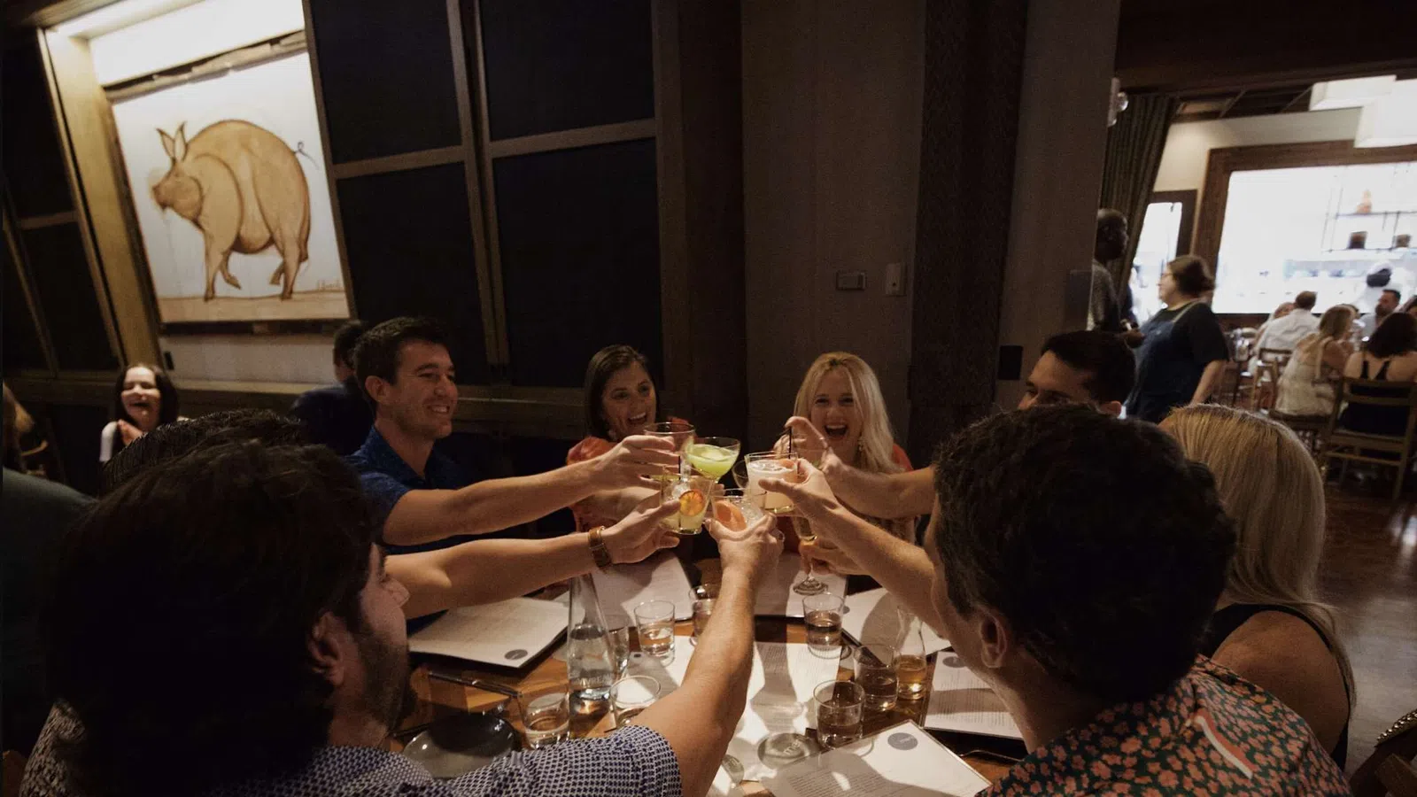 Group of friends raising glasses for a toast around a restaurant table with menus and drinks.