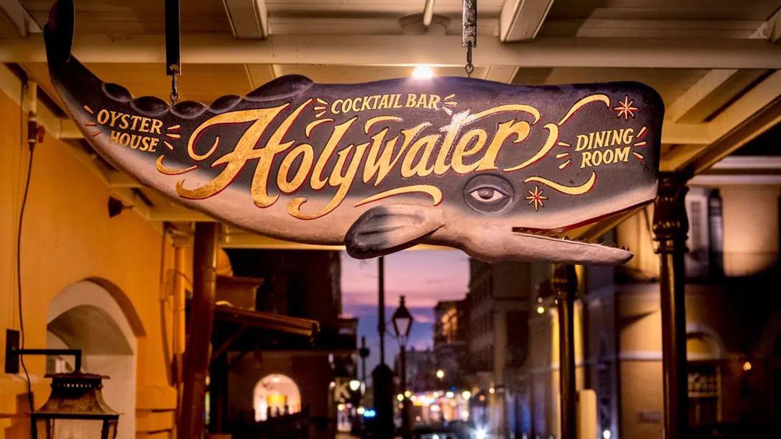 Hanging whale-shaped wooden sign for Holy Water oyster house, cocktail bar, and dining room at dusk.