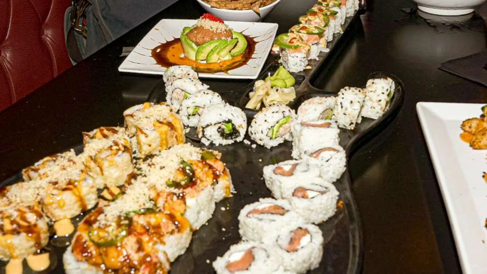 Variety of sushi rolls arranged on a black table with sauces, avocado, and garnishes in a restaurant setting.