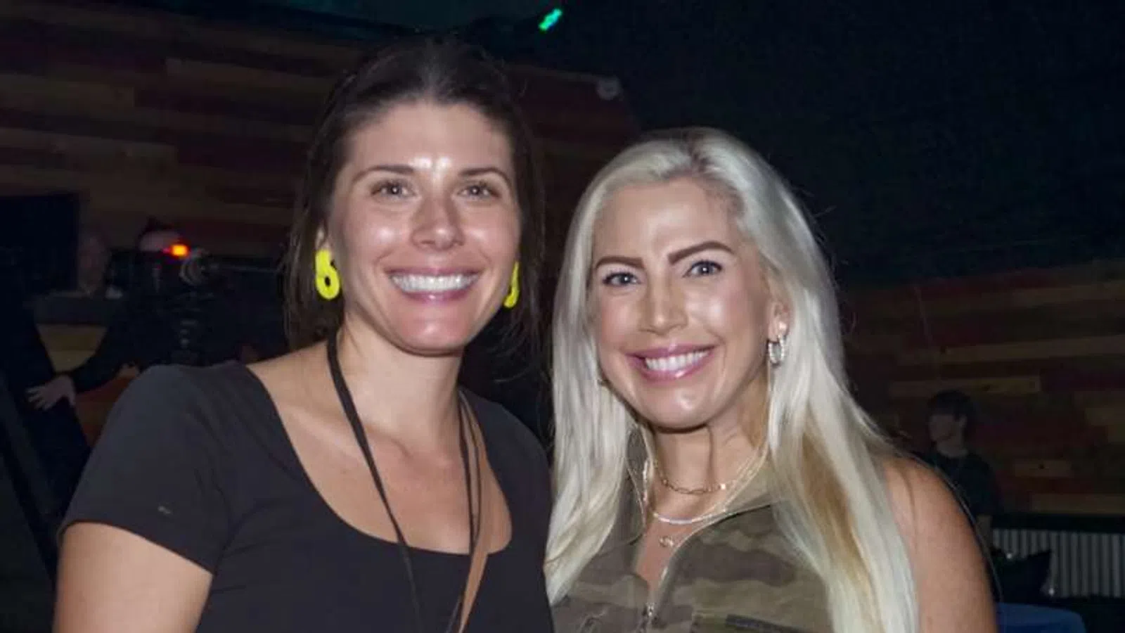 Two smiling women posing indoors, one with long blonde hair in camouflage top, the other with dark hair and yellow earrings.