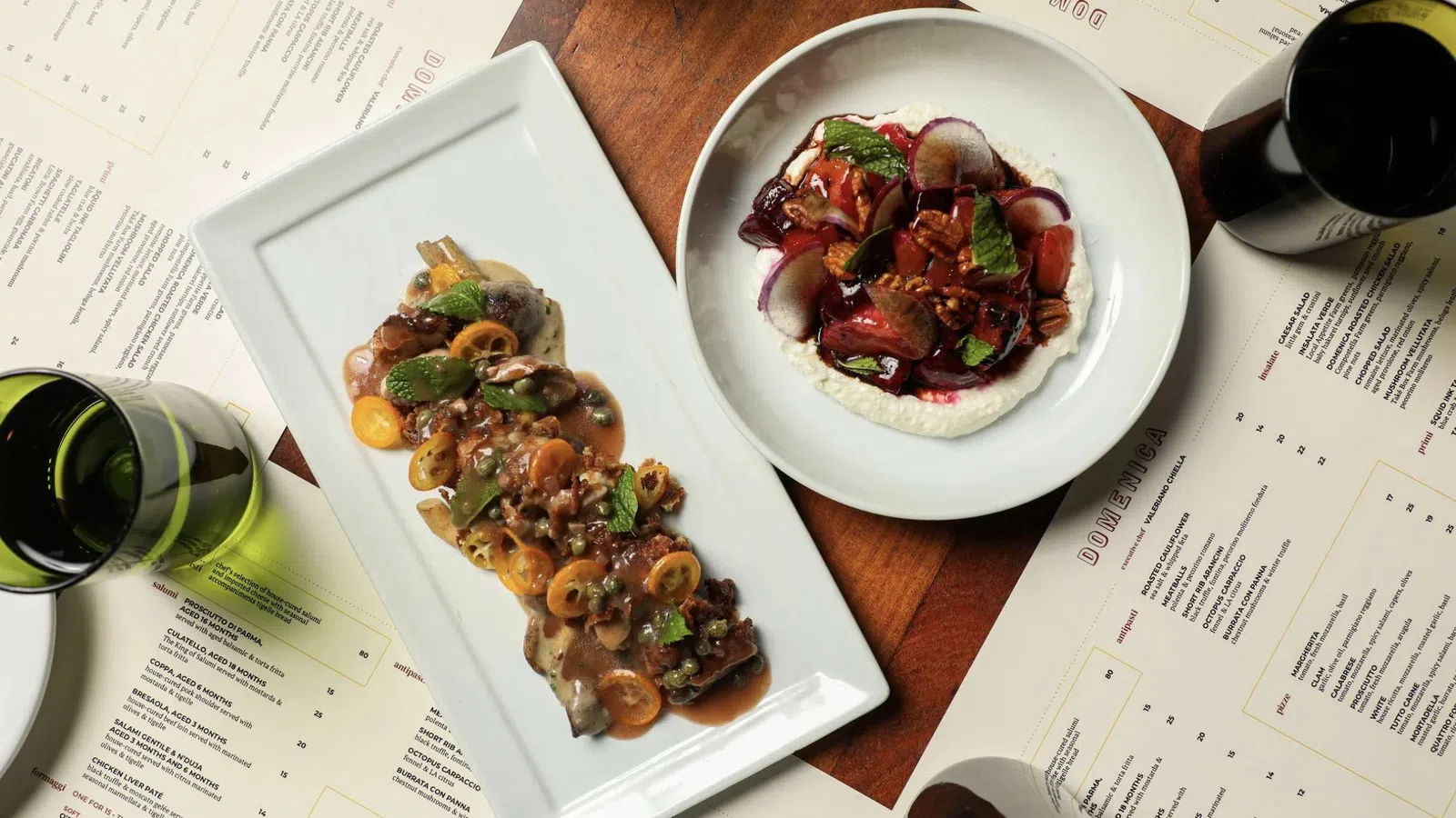 Gourmet dishes with sauces and herbs served on white plates surrounded by menus and wine glasses on wooden table.