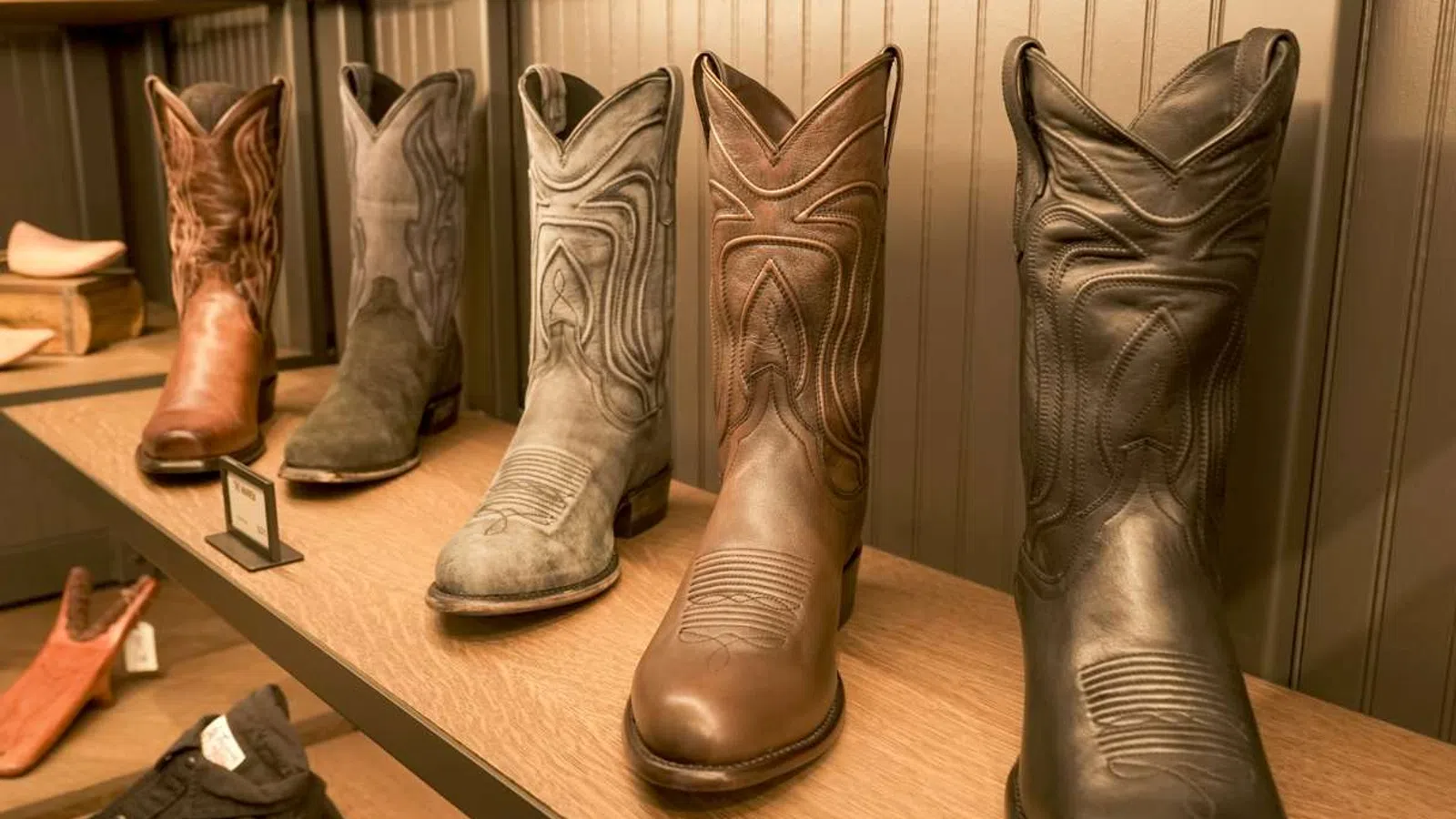 Row of six cowboy boots in various shades displayed on a wooden shelf against a paneled wall.