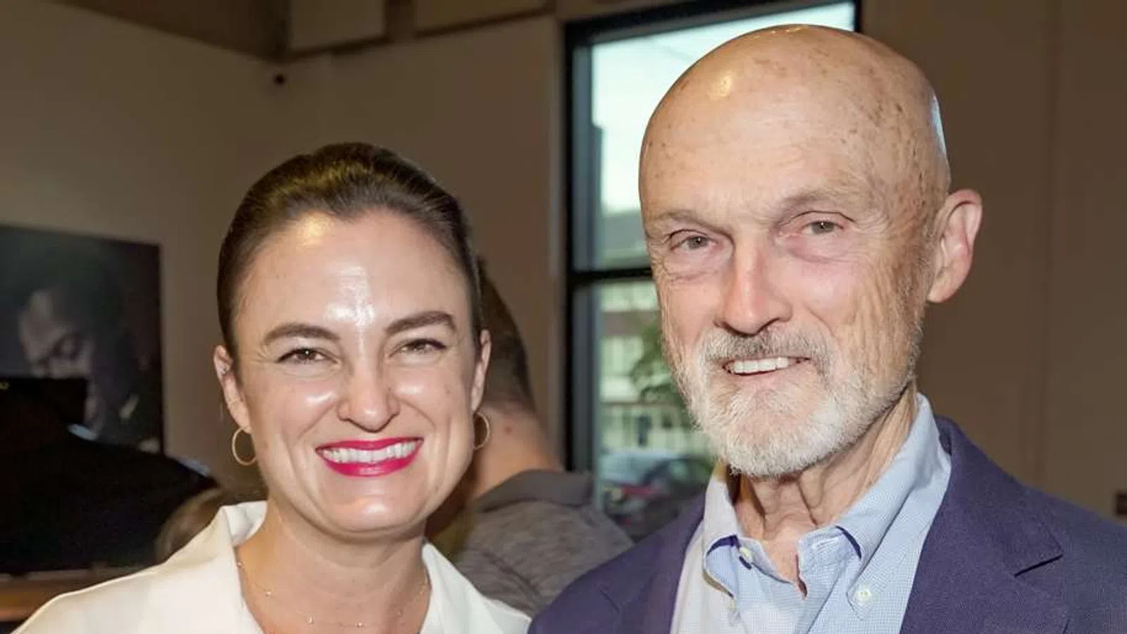 Smiling woman and older man standing indoors near a window in a casual social setting.