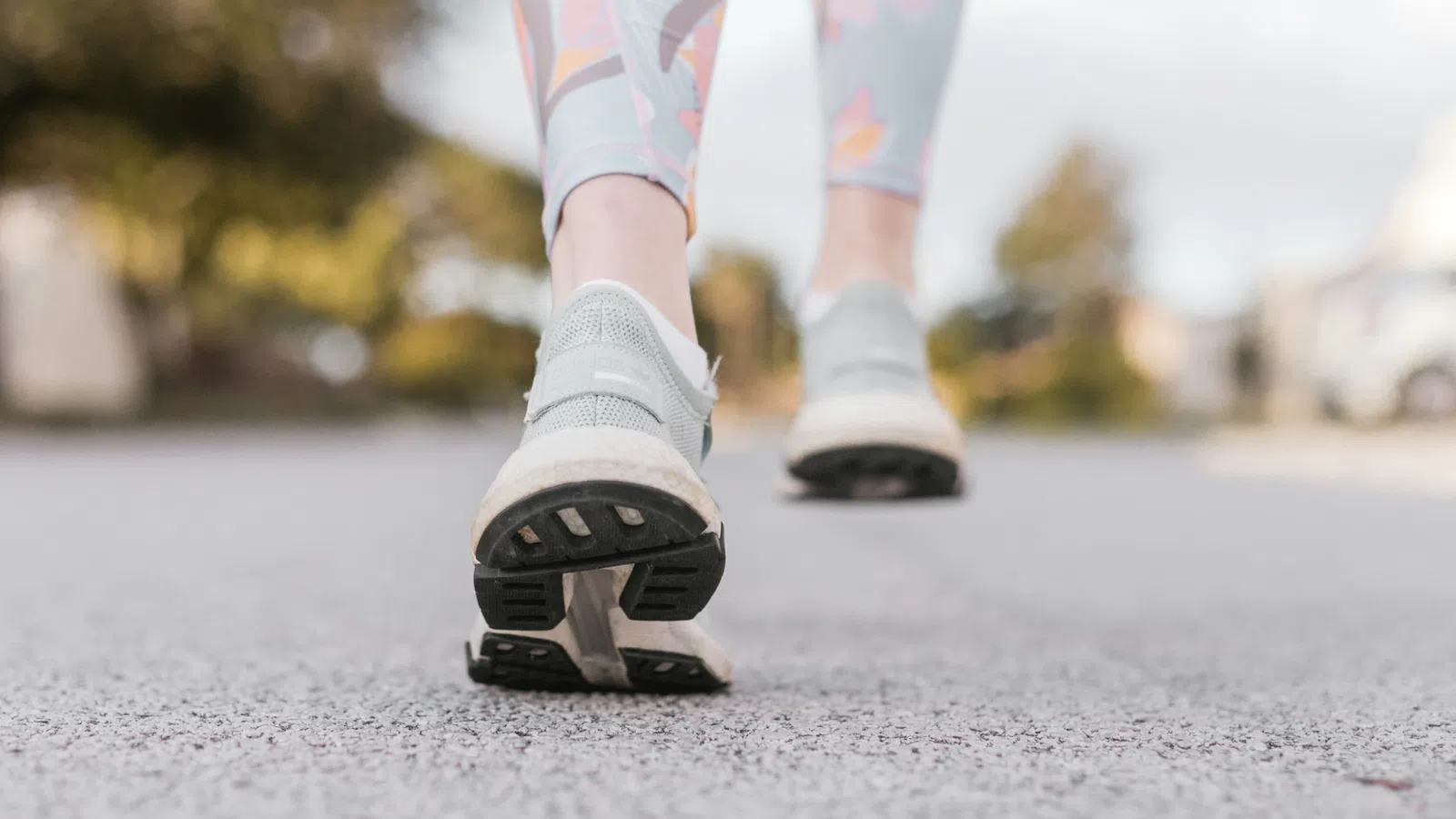 Close-up of a person walking on pavement wearing gray running shoes and floral leggings outdoors.