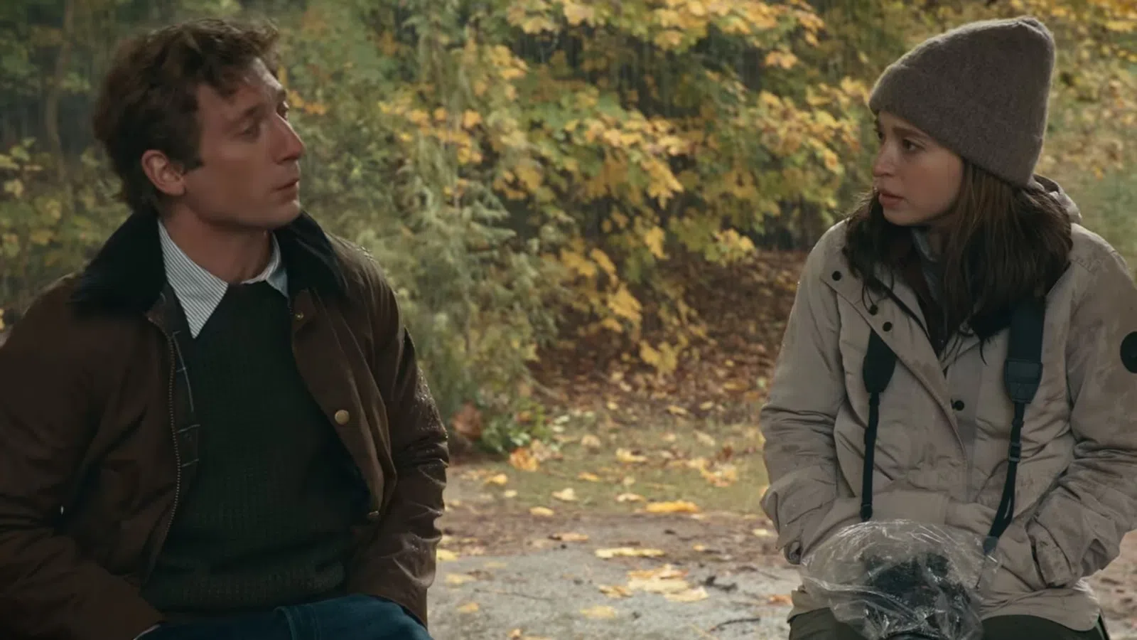 Man and woman dressed in autumn clothing sit outdoors surrounded by fall foliage during a conversation.