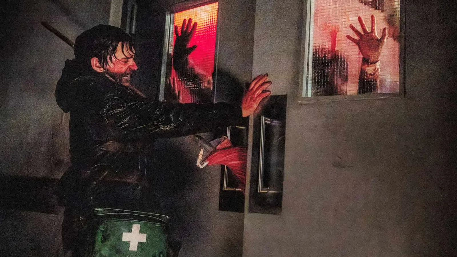 Man pushes hospital doors with bloody hands as silhouettes press against red-lit windows in a tense scene.