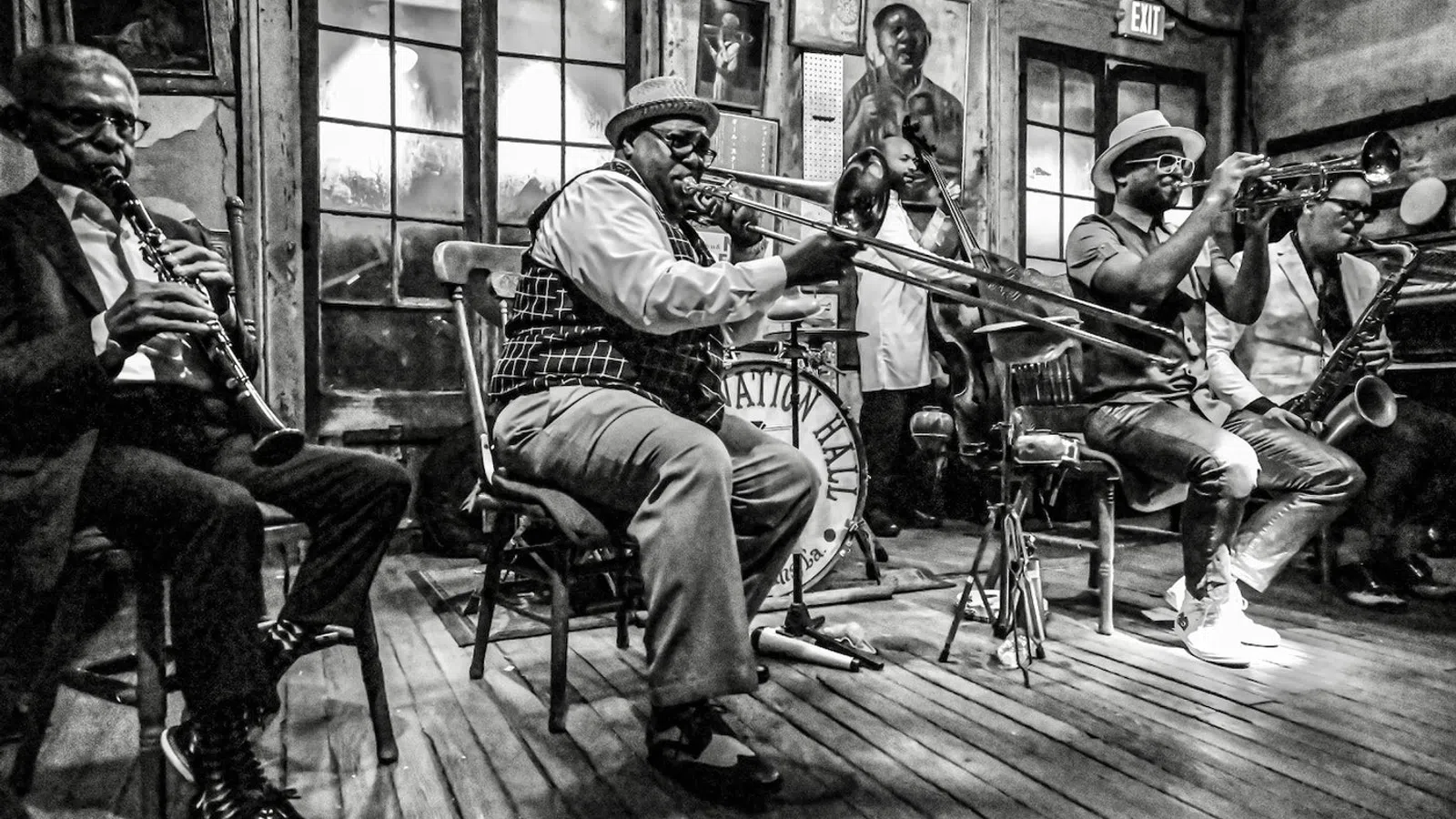 Black and white photo of jazz musicians playing saxophone, trombone, and trumpet on wooden floor stage indoors.