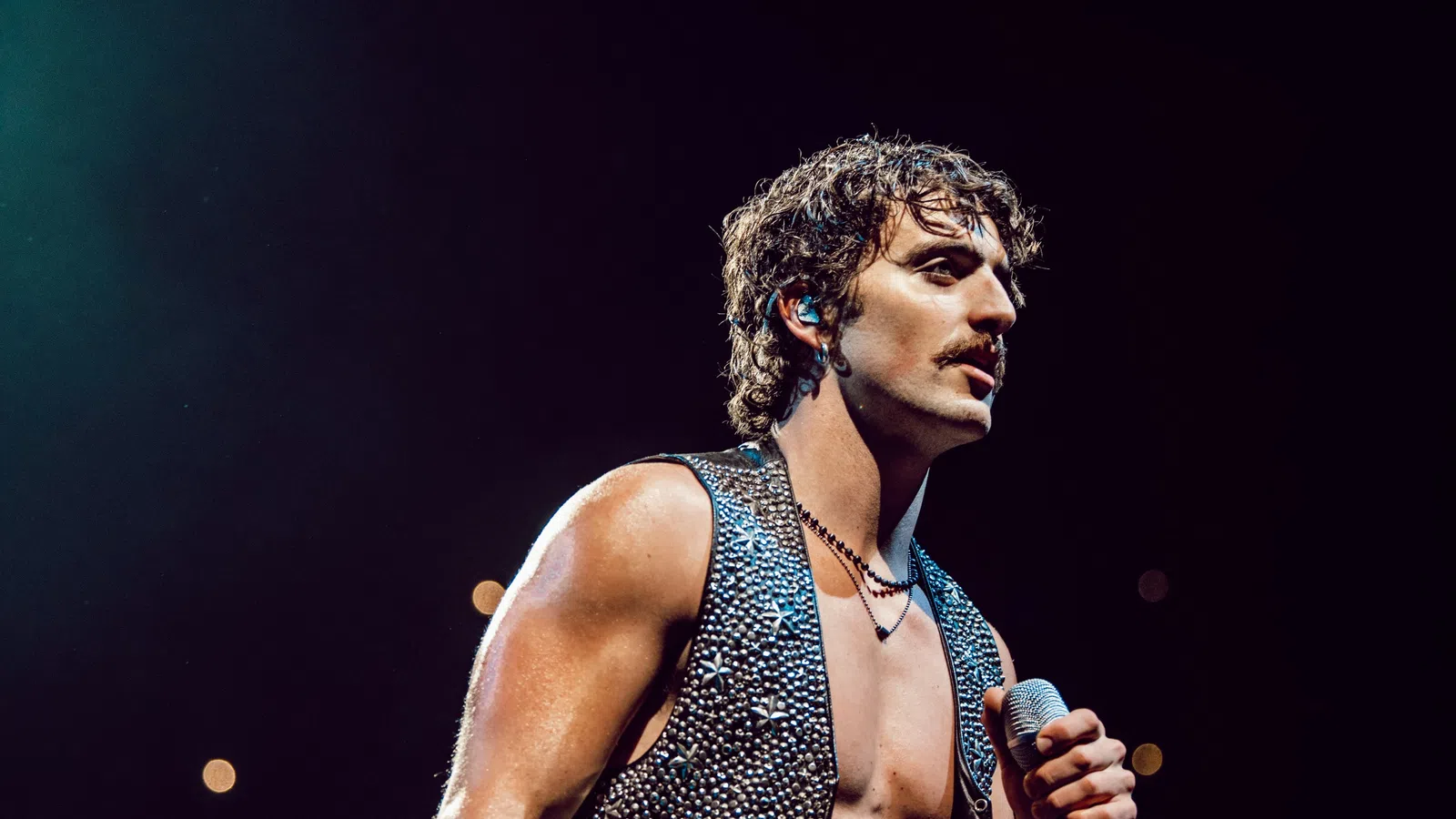 Muscular male singer with curly hair and sleeveless studded vest performing on stage with microphone