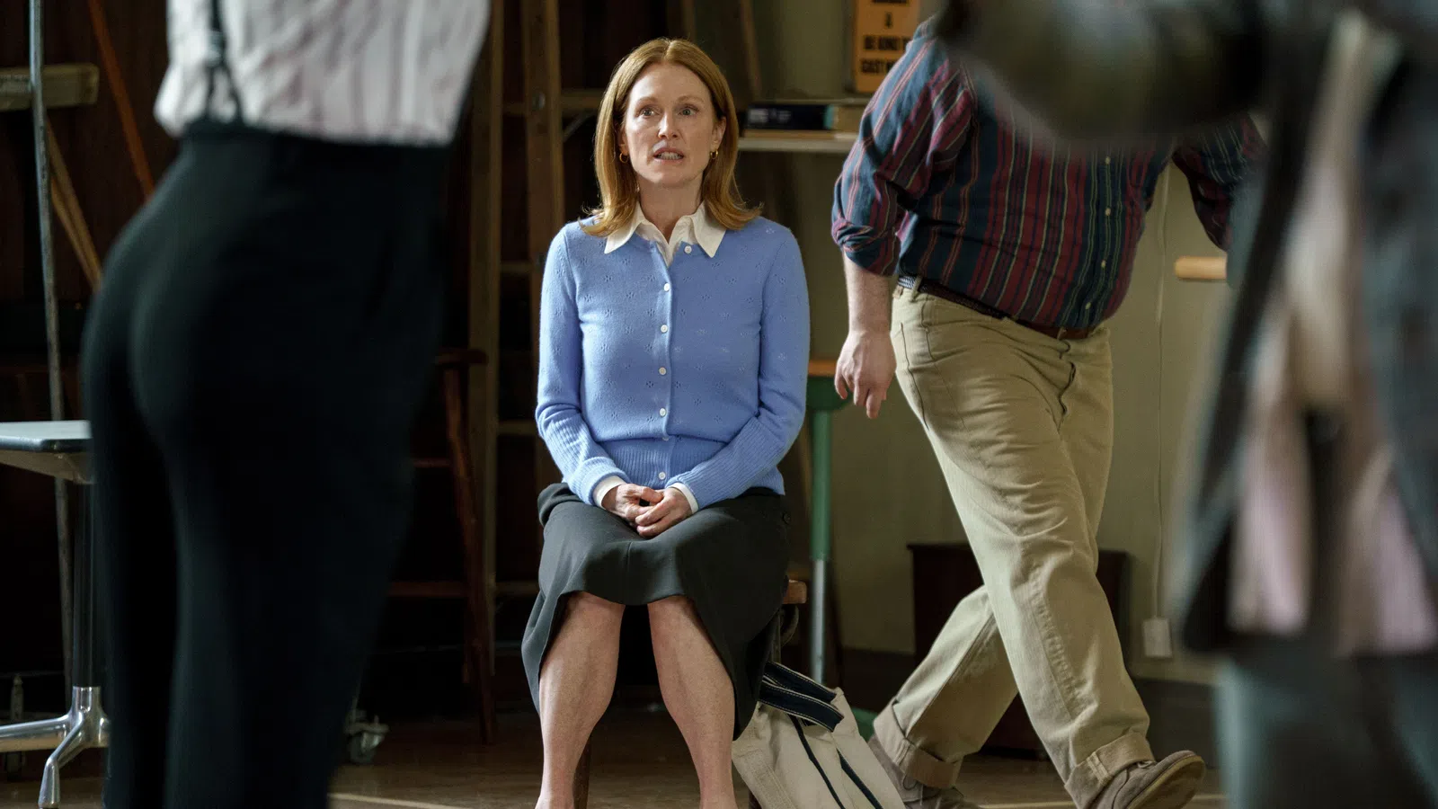 Woman in blue cardigan and black skirt sitting on a chair surrounded by blurred walking people indoors