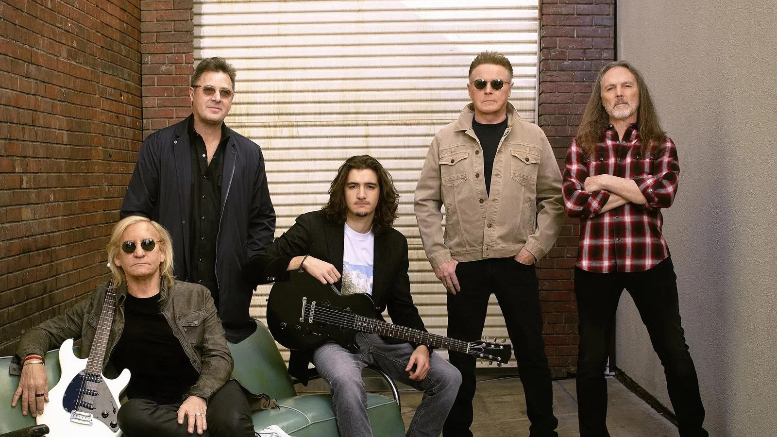 Five male musicians posing with electric guitars in an urban alley with brick walls and a closed metal door.