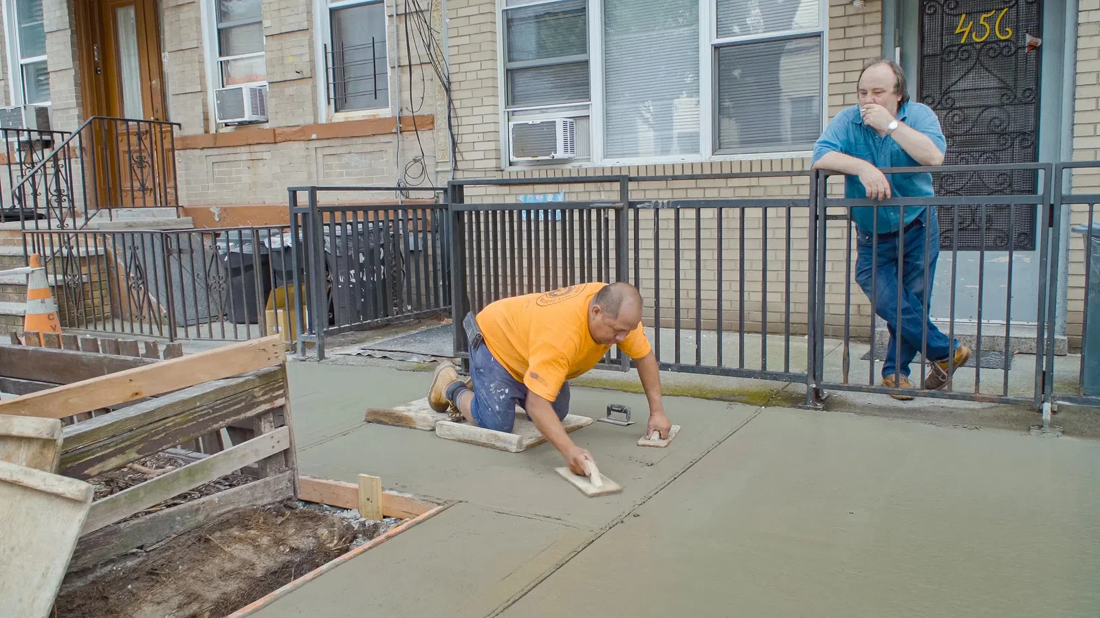 Construction worker smoothing fresh concrete on a sidewalk while another man watches at a residential building.