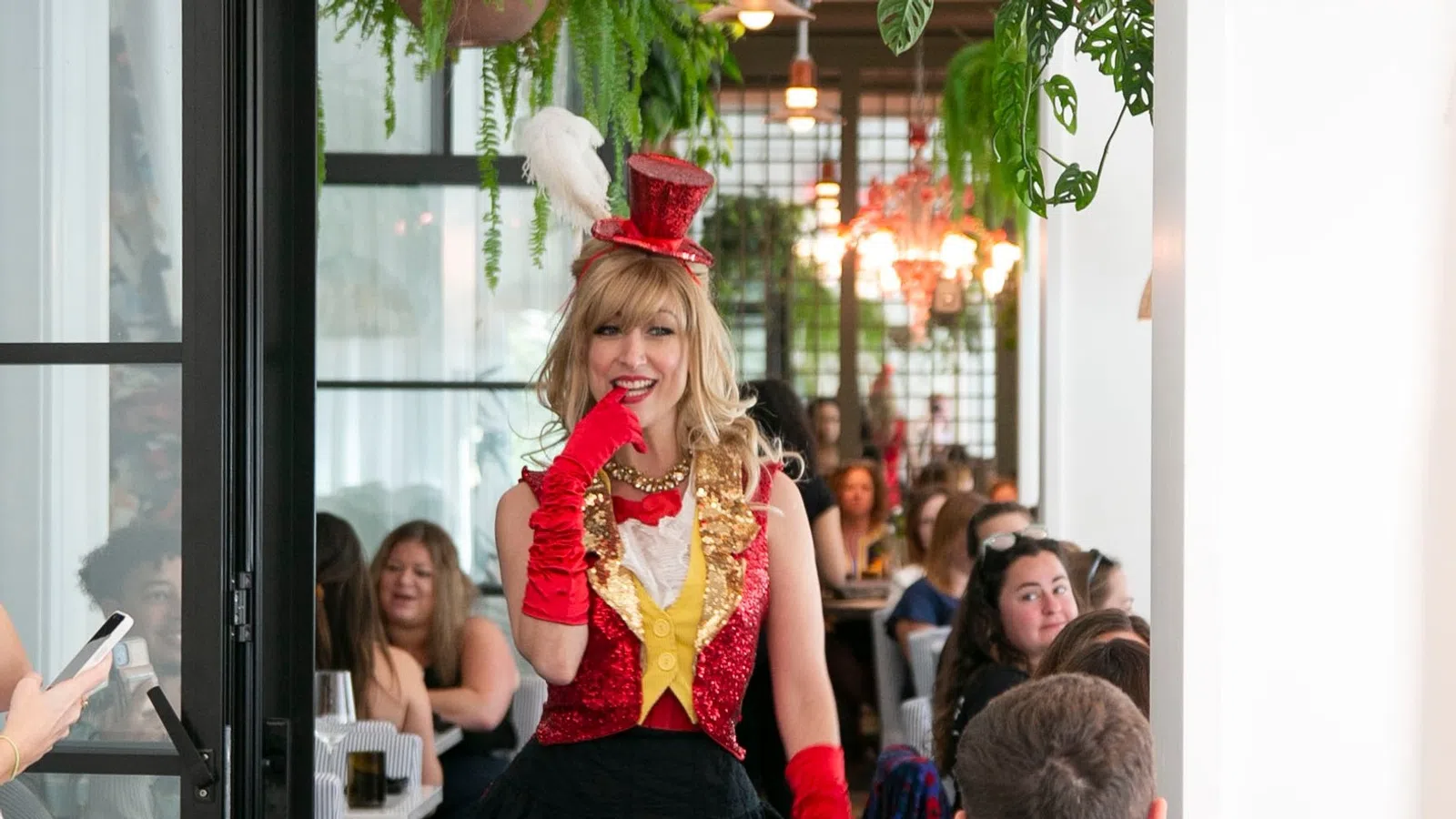 Woman in red sequined vest and hat with feather poses playfully at crowded restaurant event.