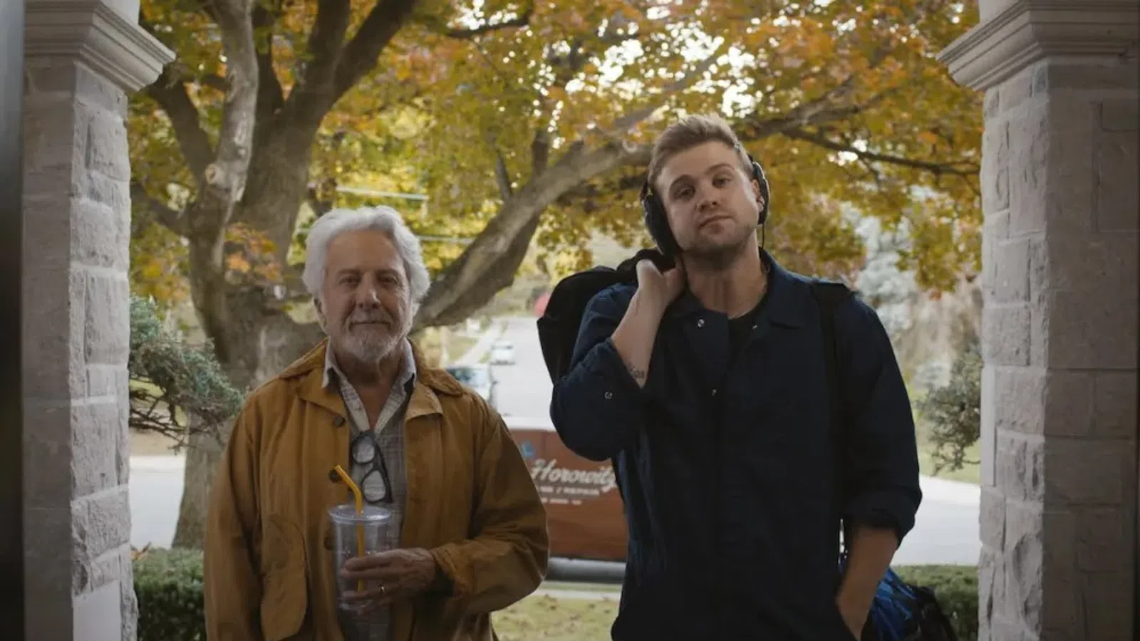 Older man with drink and younger man with headphones and backpack standing on porch in autumn setting