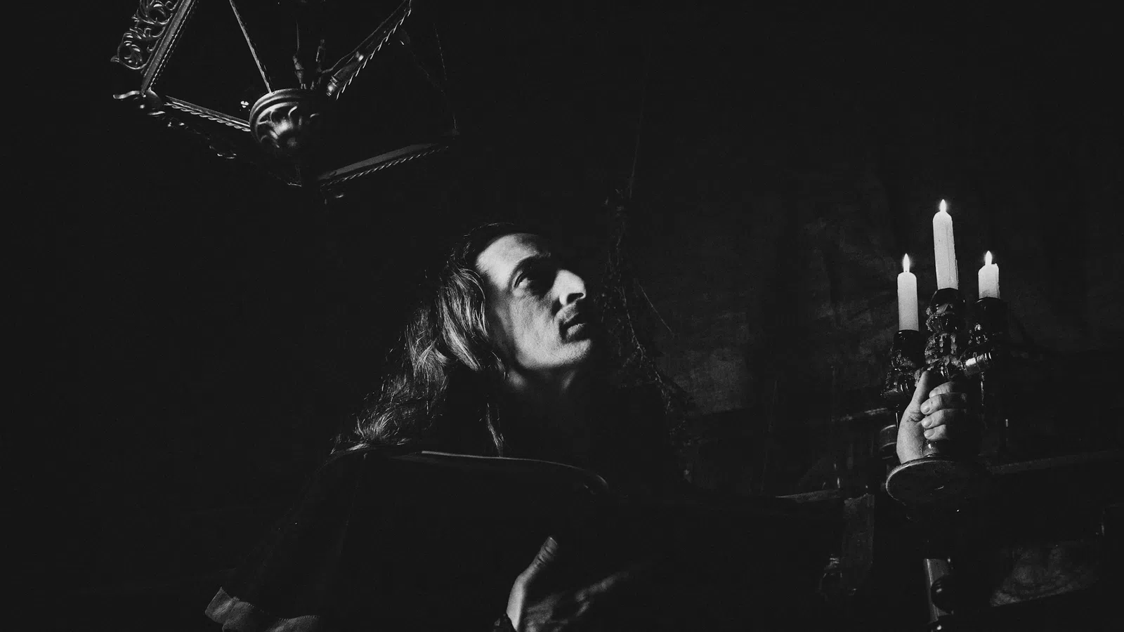 Black and white photo of a person holding a candelabra with burning candles in a dark, gothic setting.