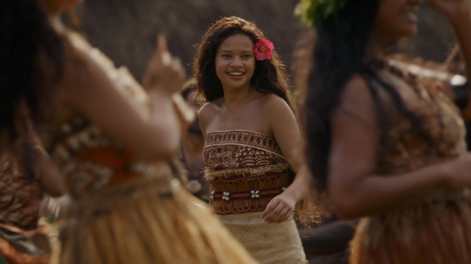 Smiling woman with a flower in her hair dances in traditional Polynesian attire among a group of dancers.